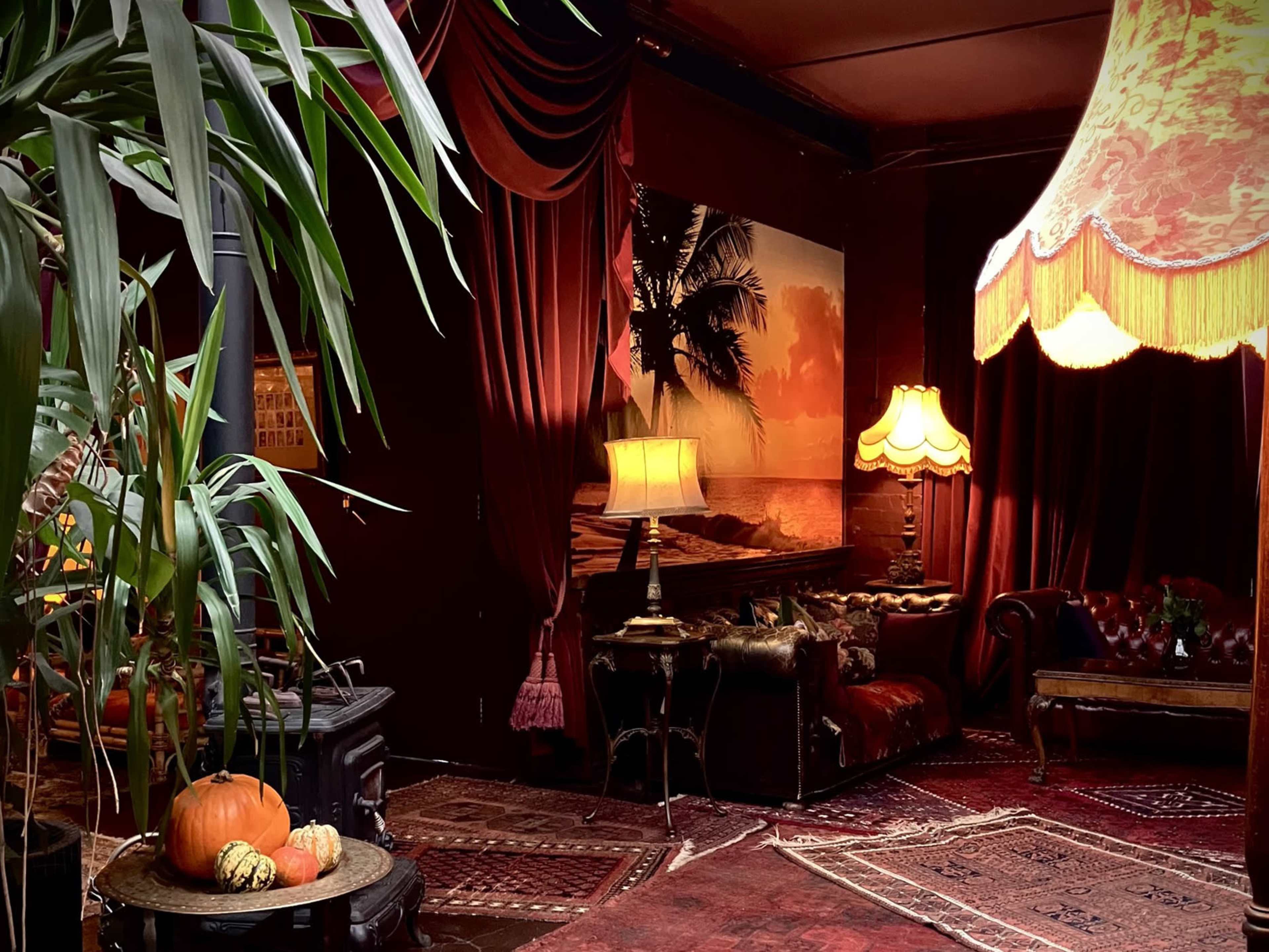 A warmly lit, vintage-style lounge featuring a palm tree mural, leather furniture, and decorative lamps, accented by greenery and autumnal decor.
