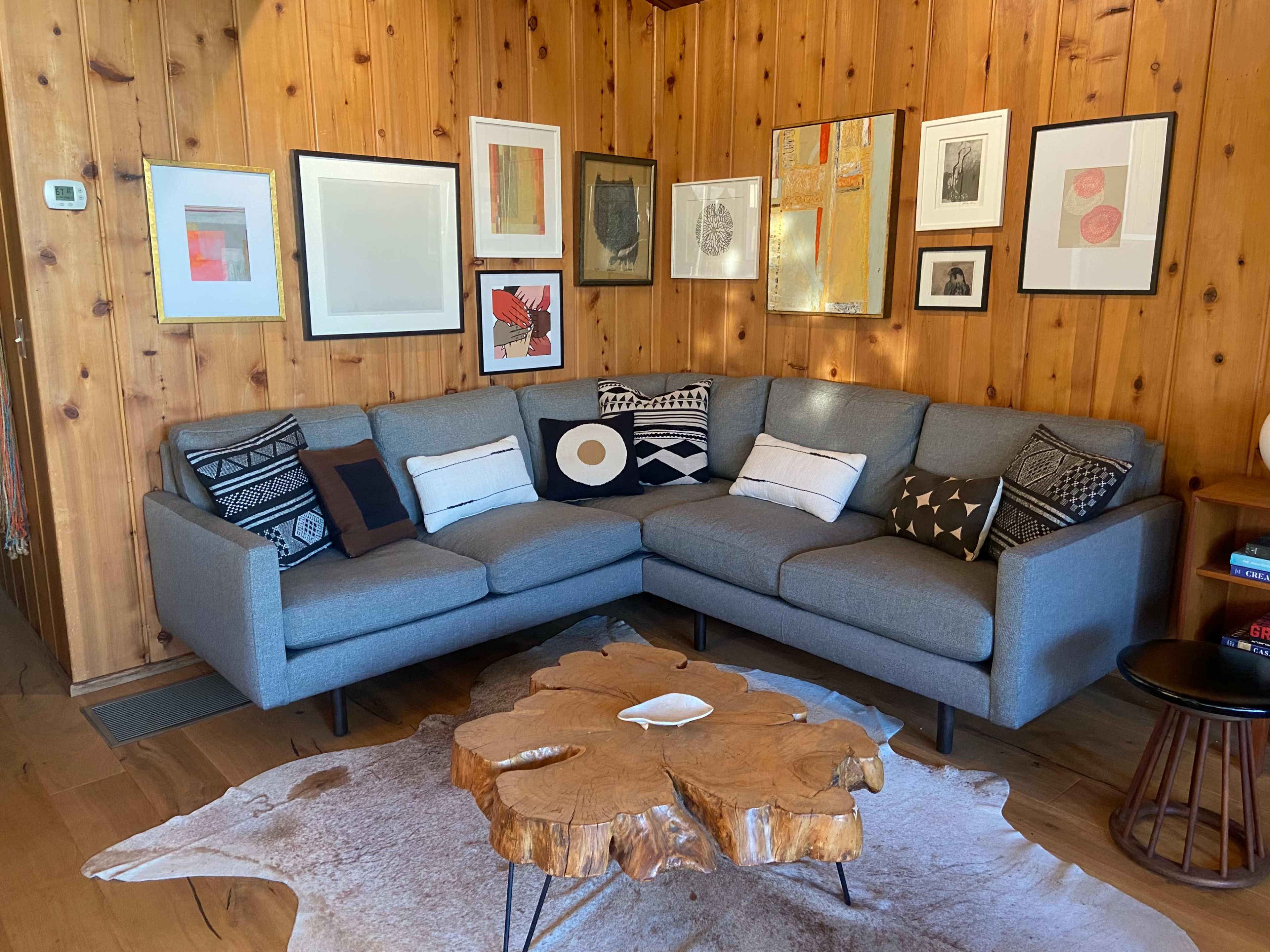 The scene features a cozy living room with a gray sectional sofa, a wooden coffee table, and various framed artworks on the wooden paneled walls.