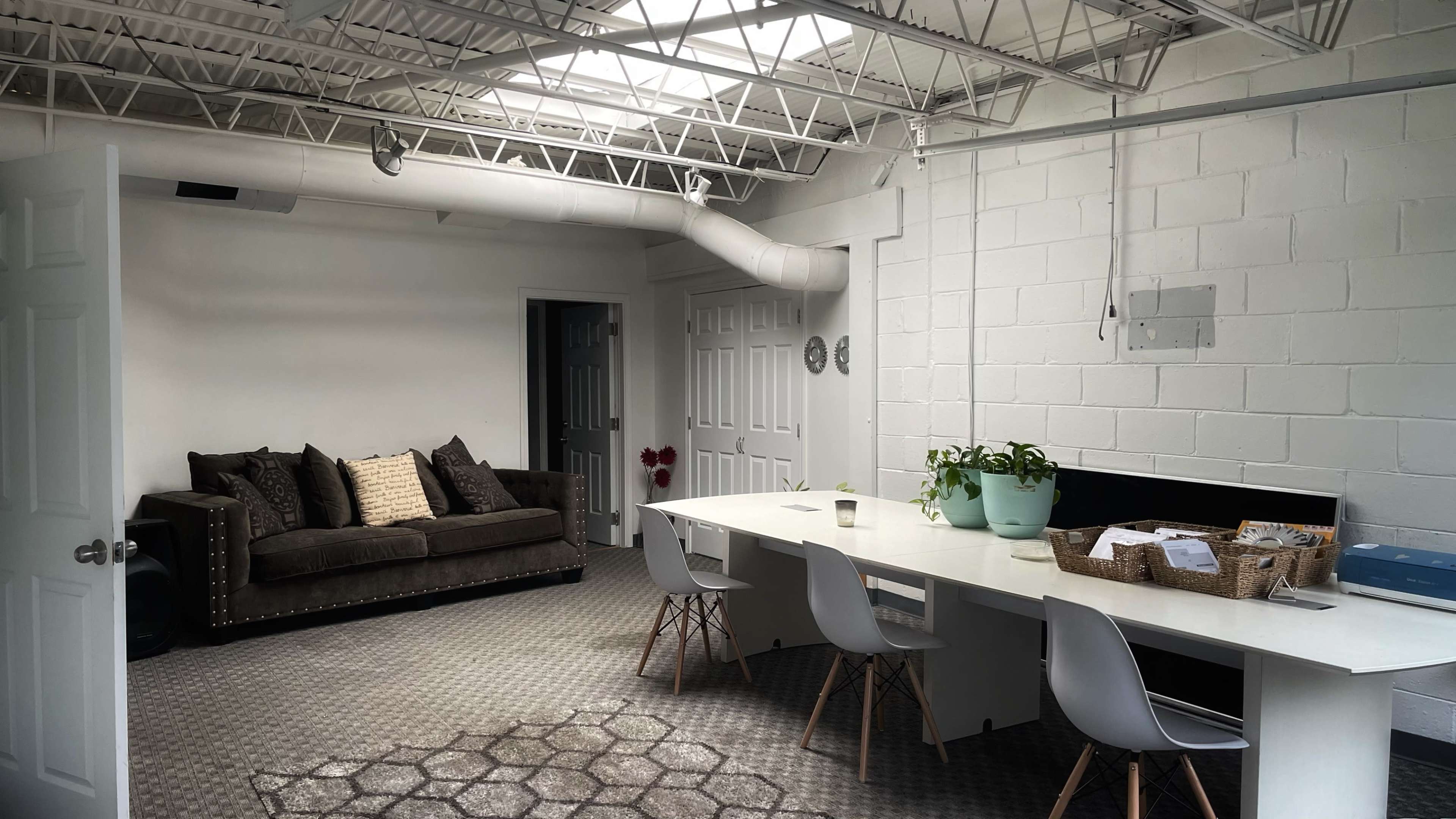 A modern office space with a sofa on one side and a long table with chairs on the other, featuring large windows and exposed ductwork overhead.