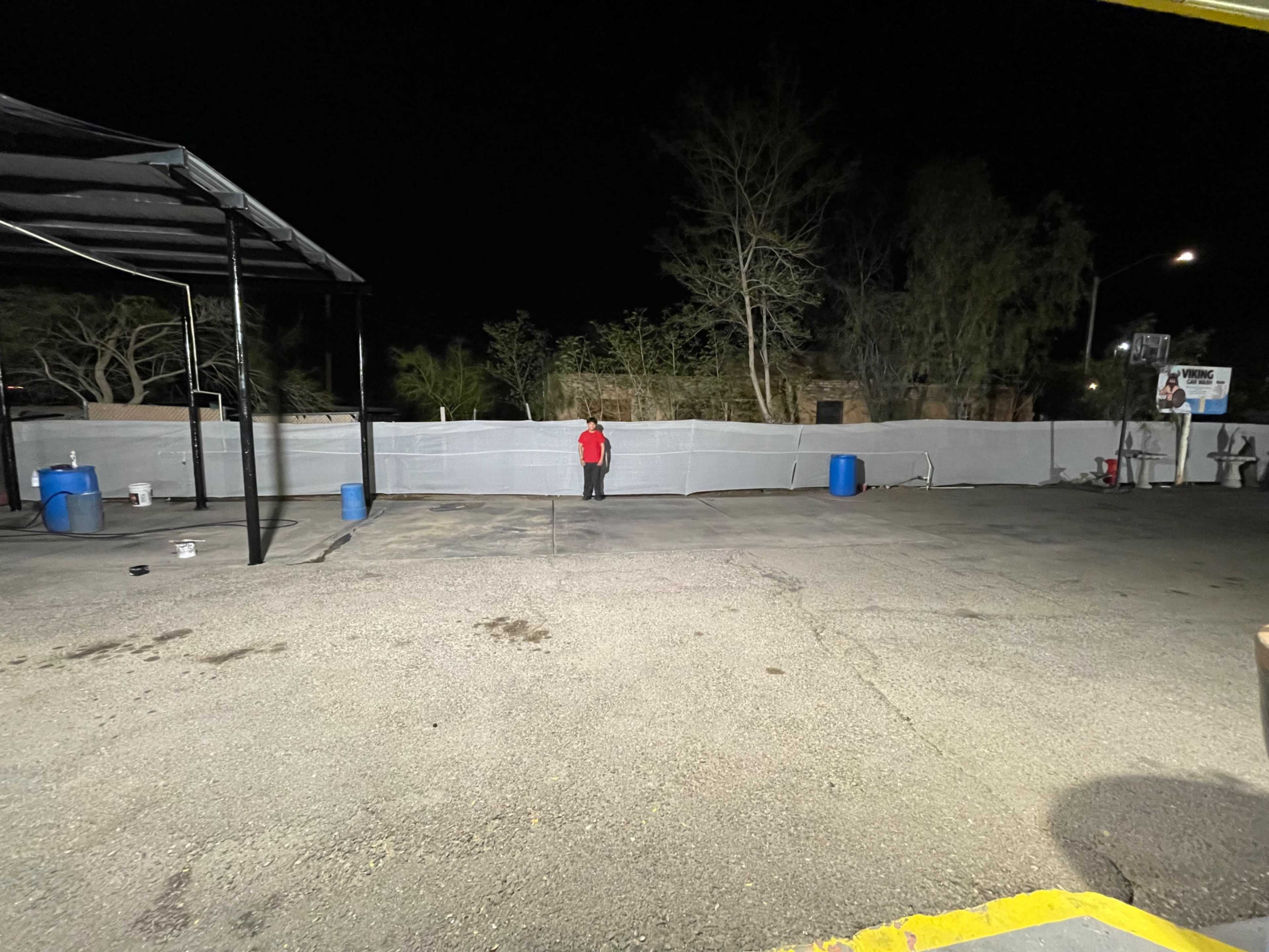 A person stands alone in a dark area next to a fenced-off section with blue barrels and an unfinished surface.