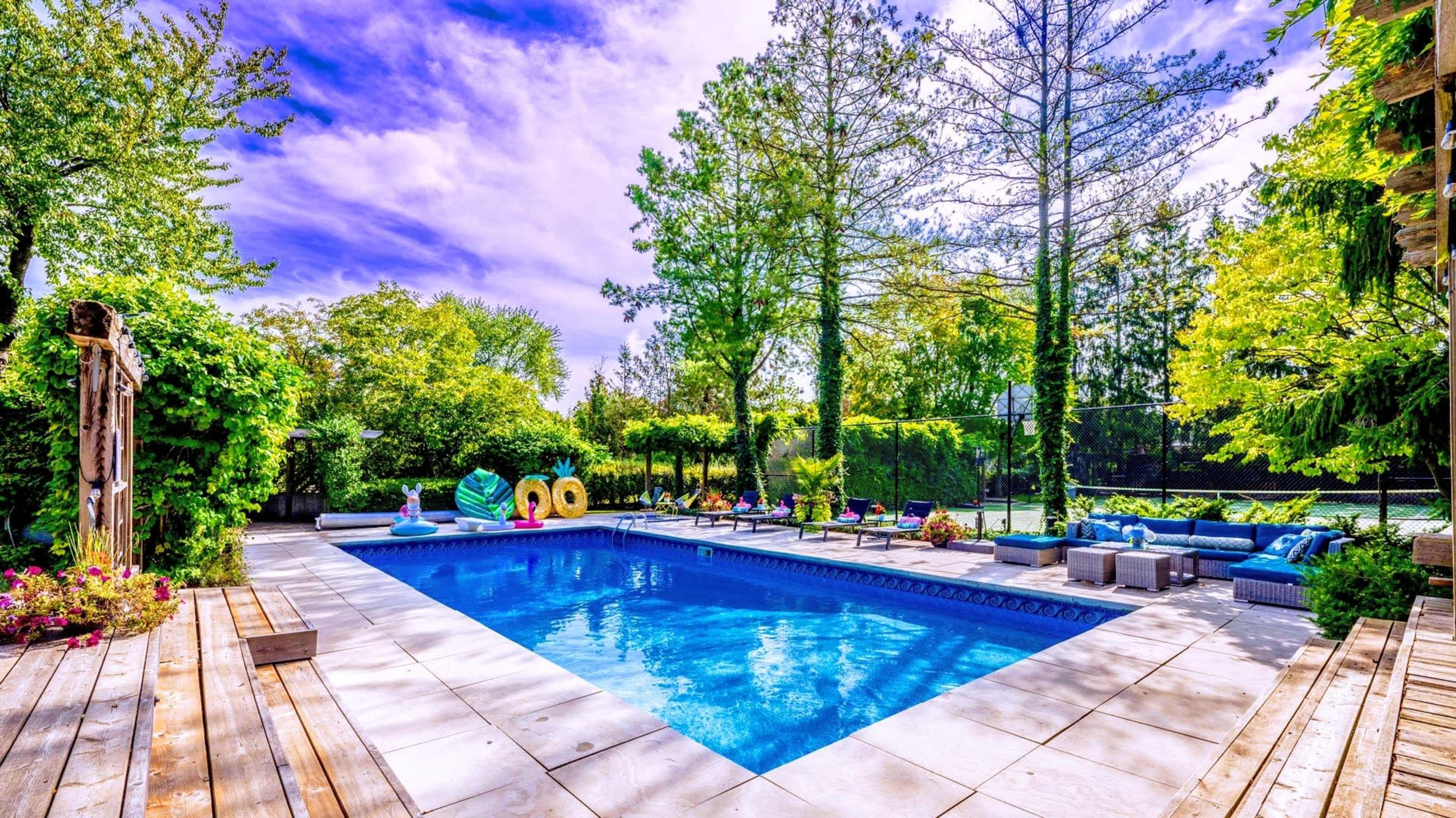 A backyard swimming pool surrounded by green trees and lounge furniture, with colorful floaties in the water.