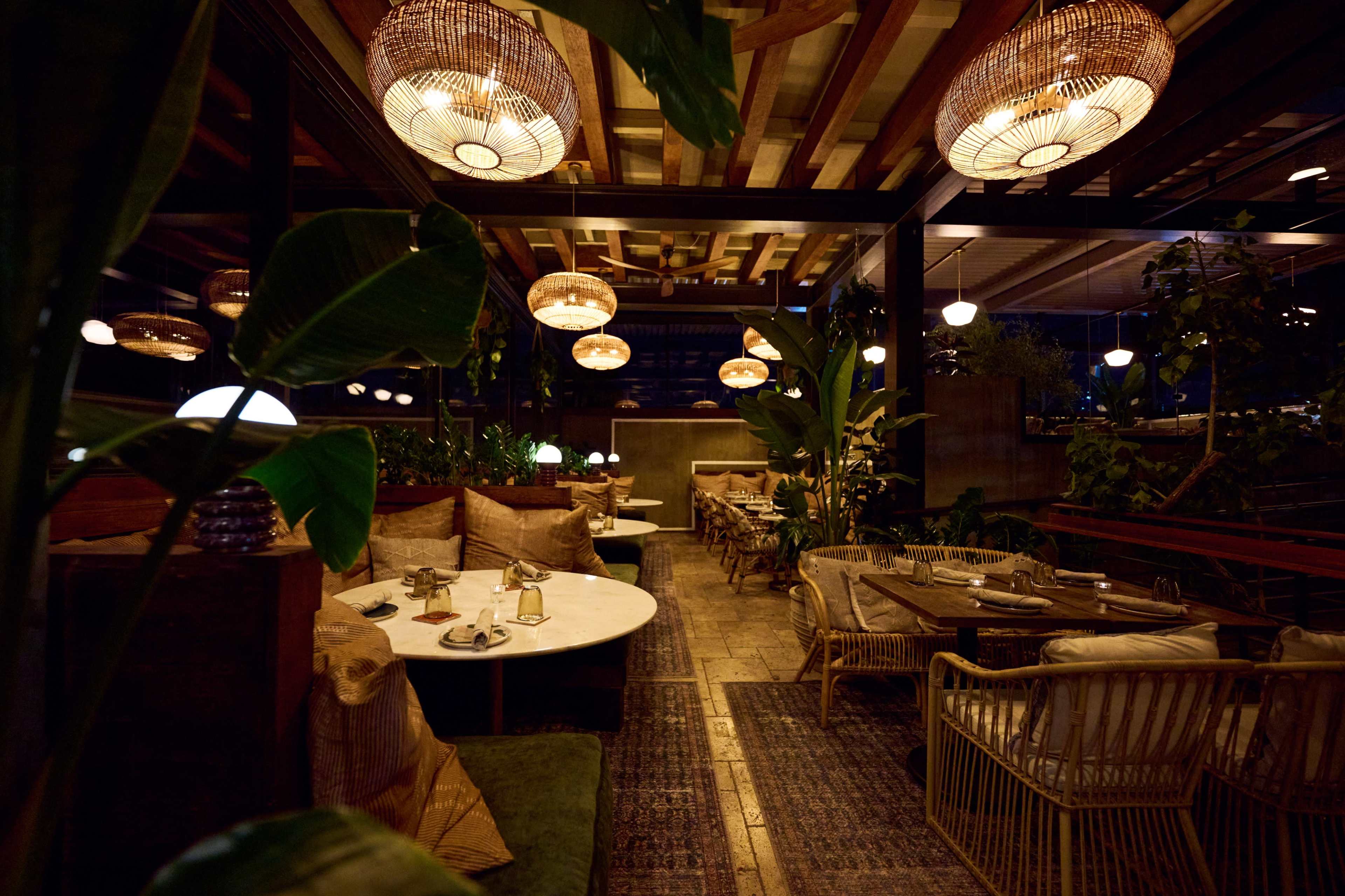 The image depicts a stylish restaurant interior with woven light fixtures, wooden beams, and a mix of seating arrangements, including tables and cozy booths surrounded by plants.