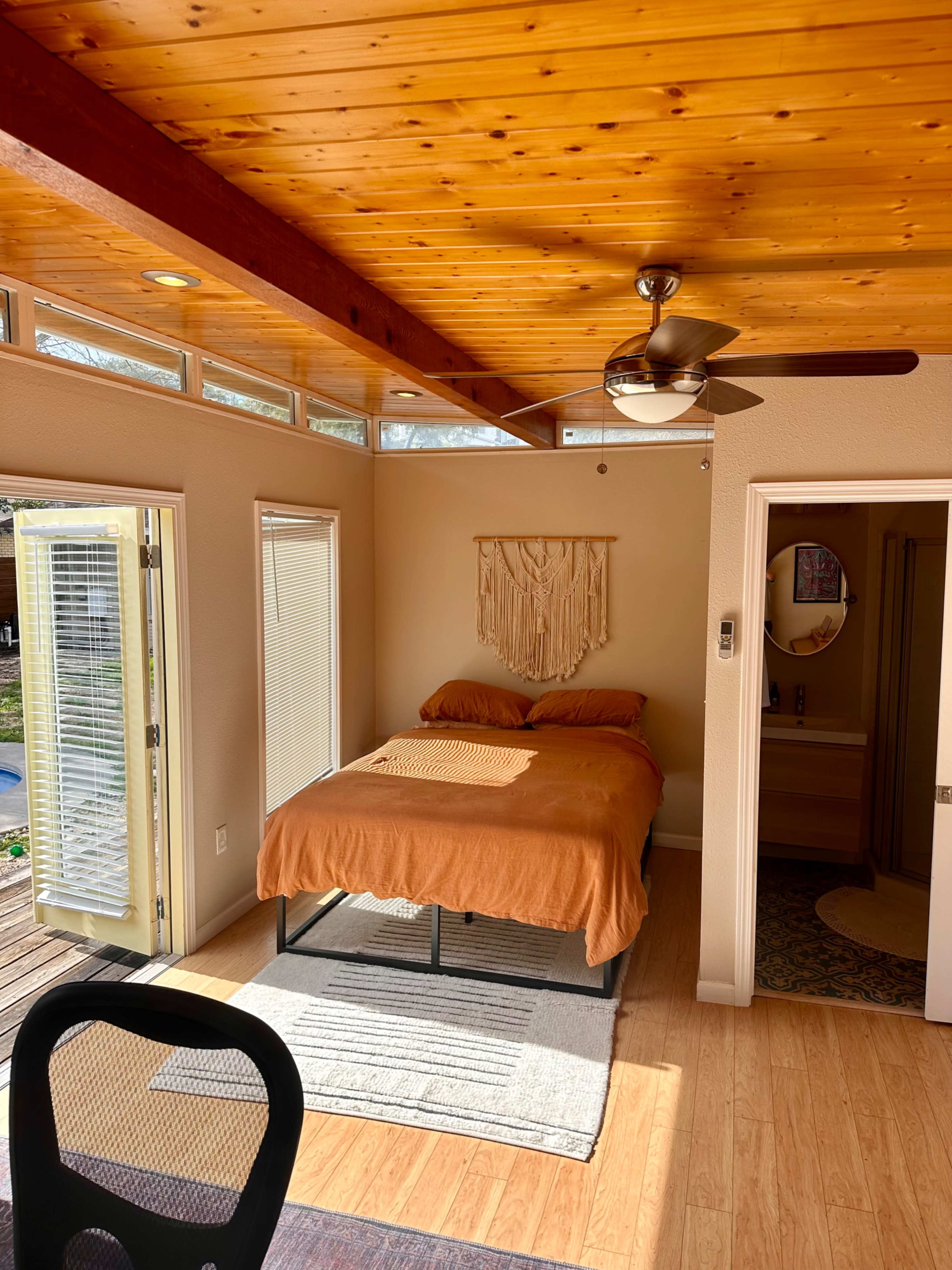 A small bedroom features a bed with an orange blanket, wooden ceiling beams, and large windows allowing natural light.
