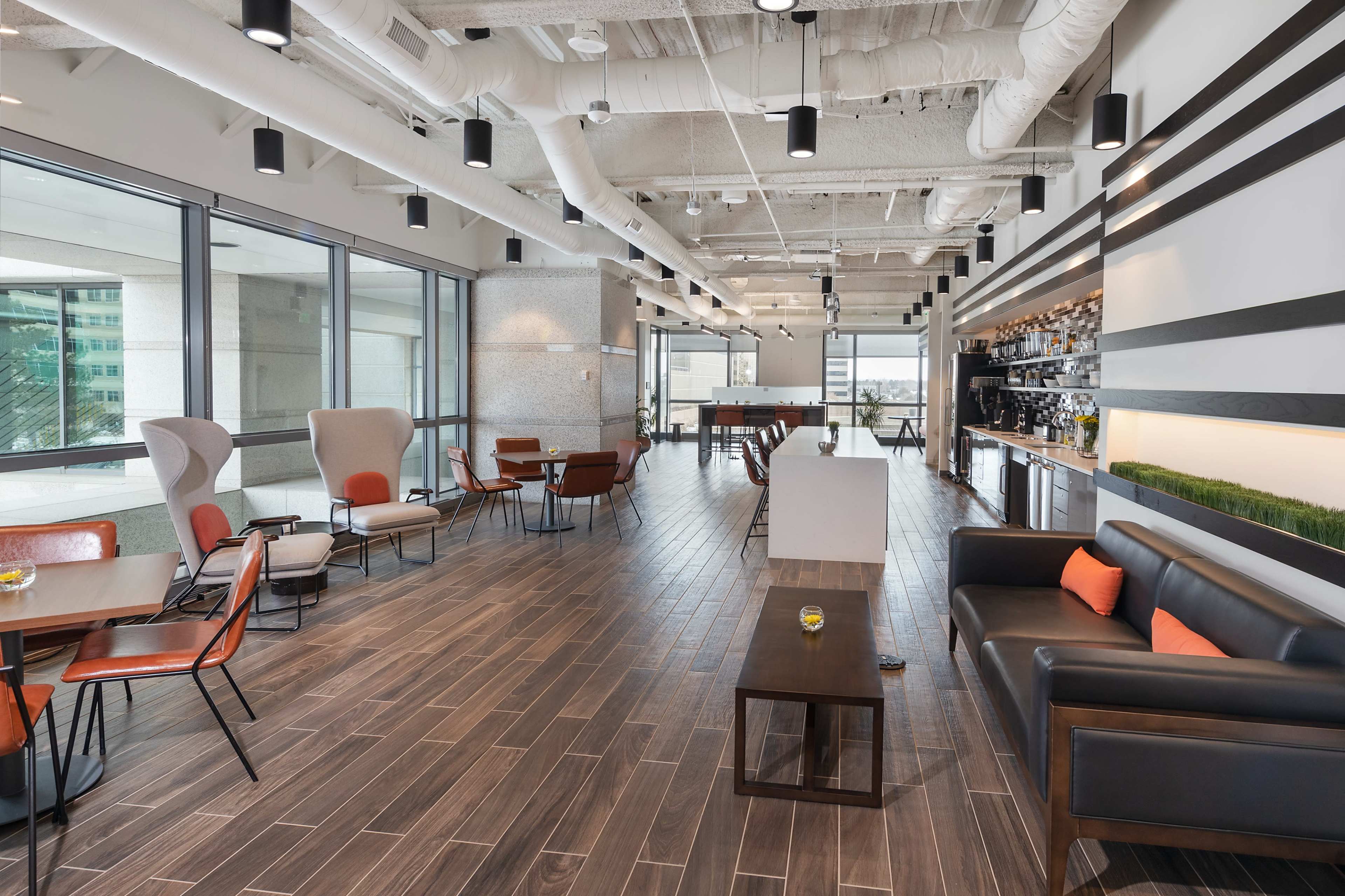 The image shows a modern office lounge area with seating arrangements, wooden flooring, and large windows providing natural light.