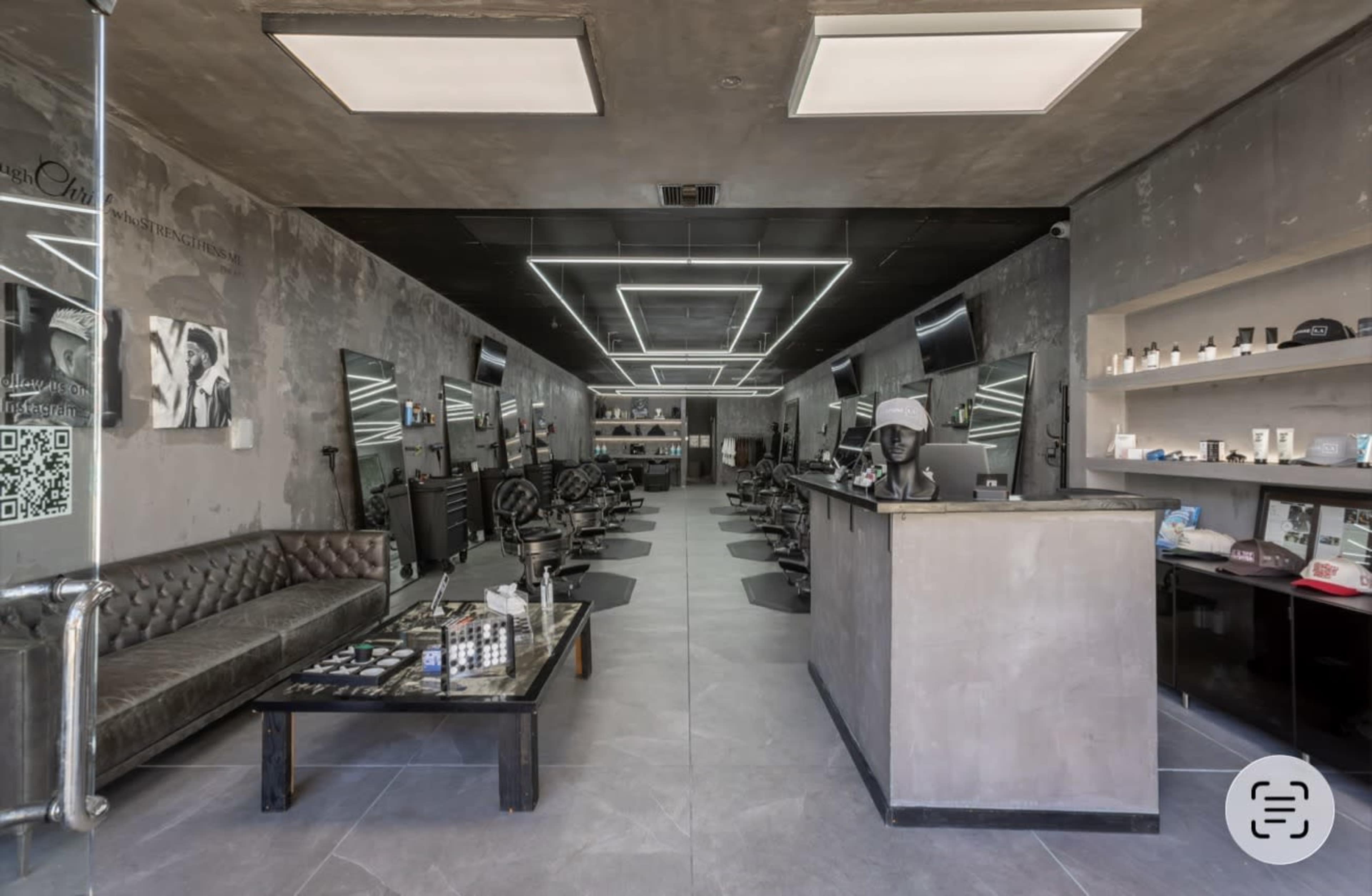 The interior of a modern barbershop features sleek black leather chairs, a long table with grooming products, and illuminated ceiling panels.
