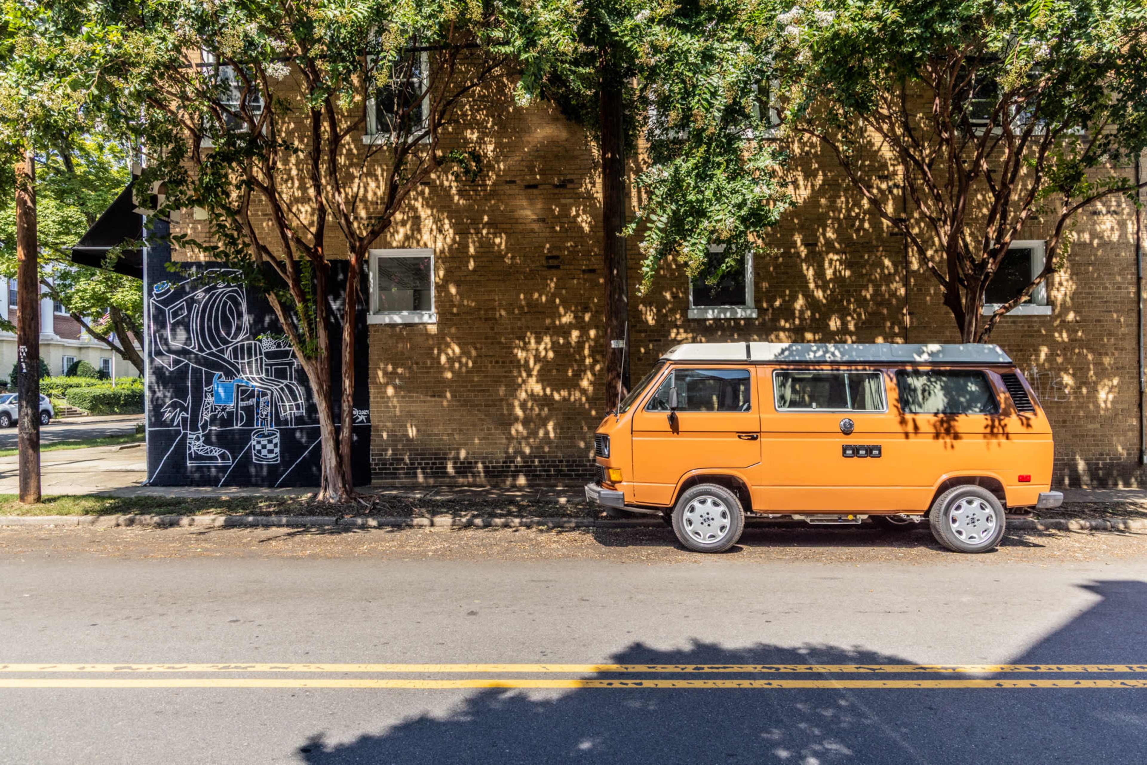 An orange vintage van is parked next to a brick building with trees casting shadows on the sidewalk.