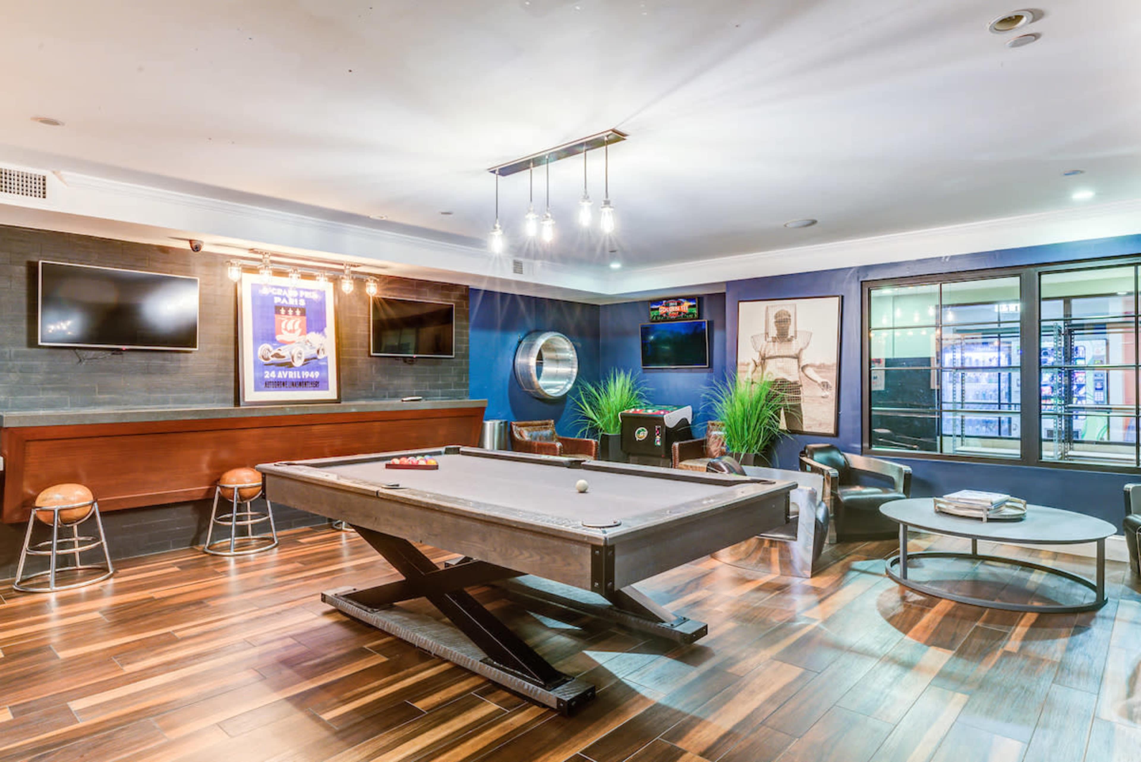 A game room featuring a billiard table, bar seating, and several television screens on the walls.