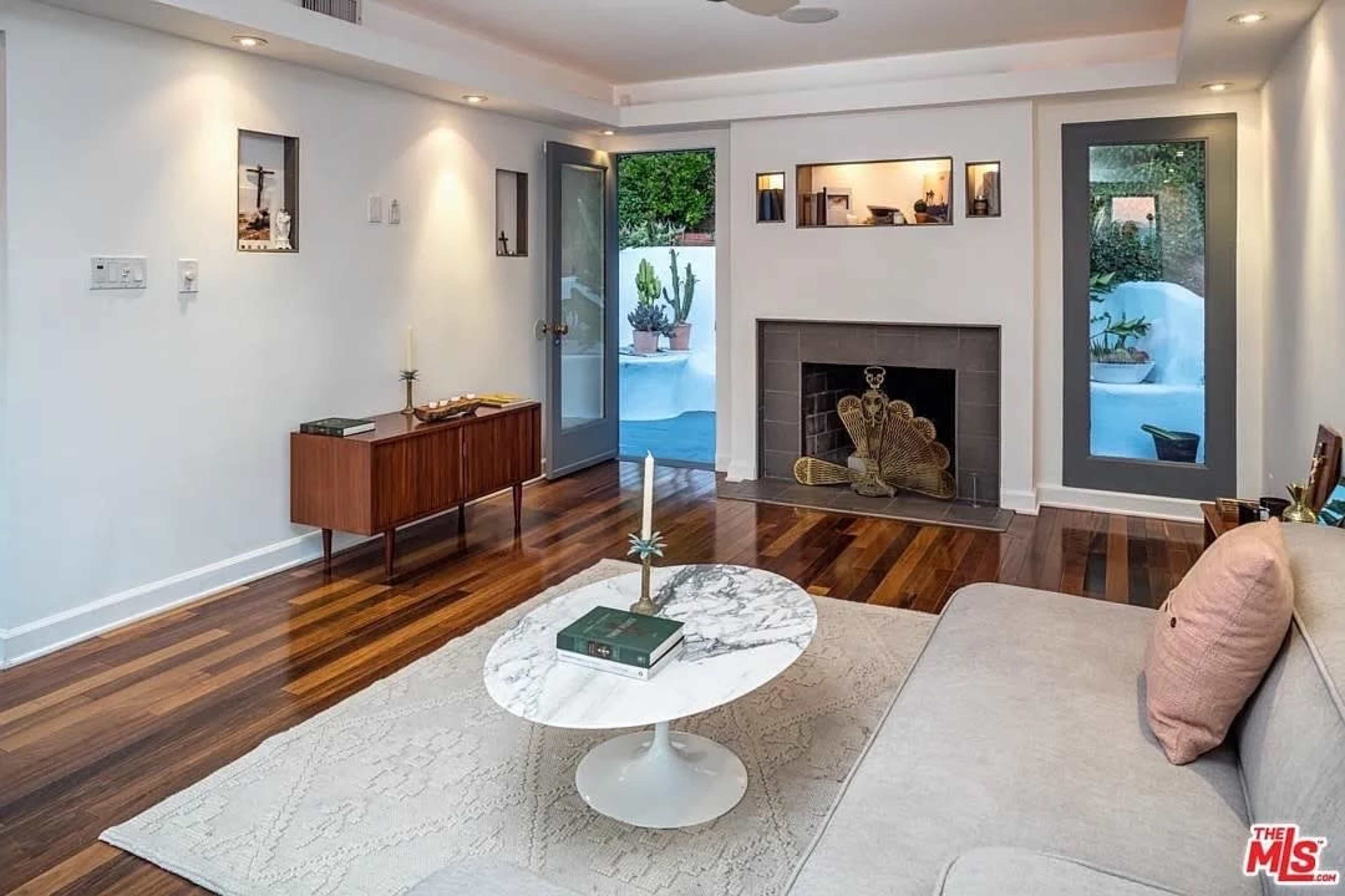 The image shows a modern living room with a marble coffee table, a light-colored sofa, a wooden side table, and a fireplace, featuring a sliding glass door that opens to an outdoor area.