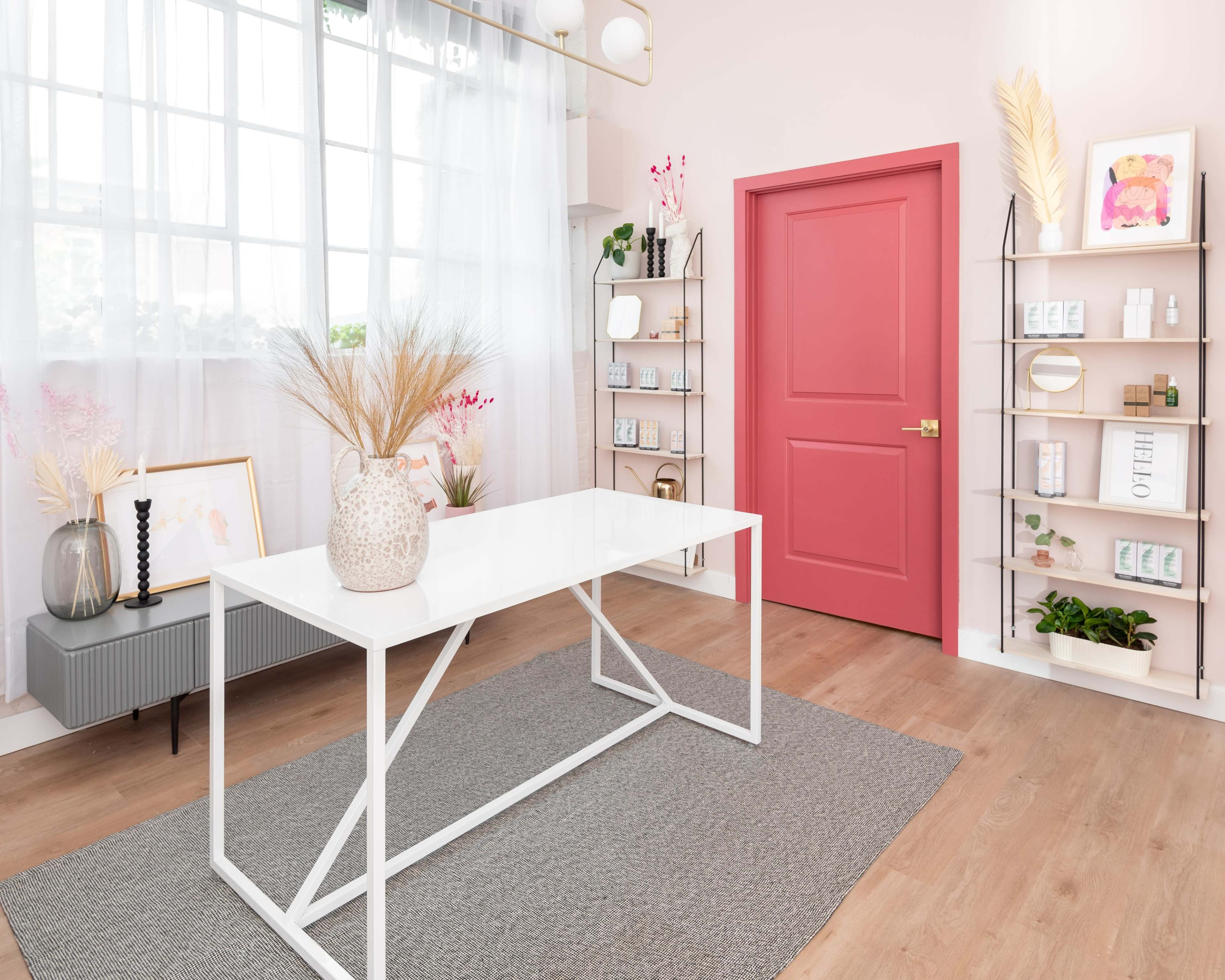 A modern room with a white table, a coral door, shelves displaying products, and decorative plants.
