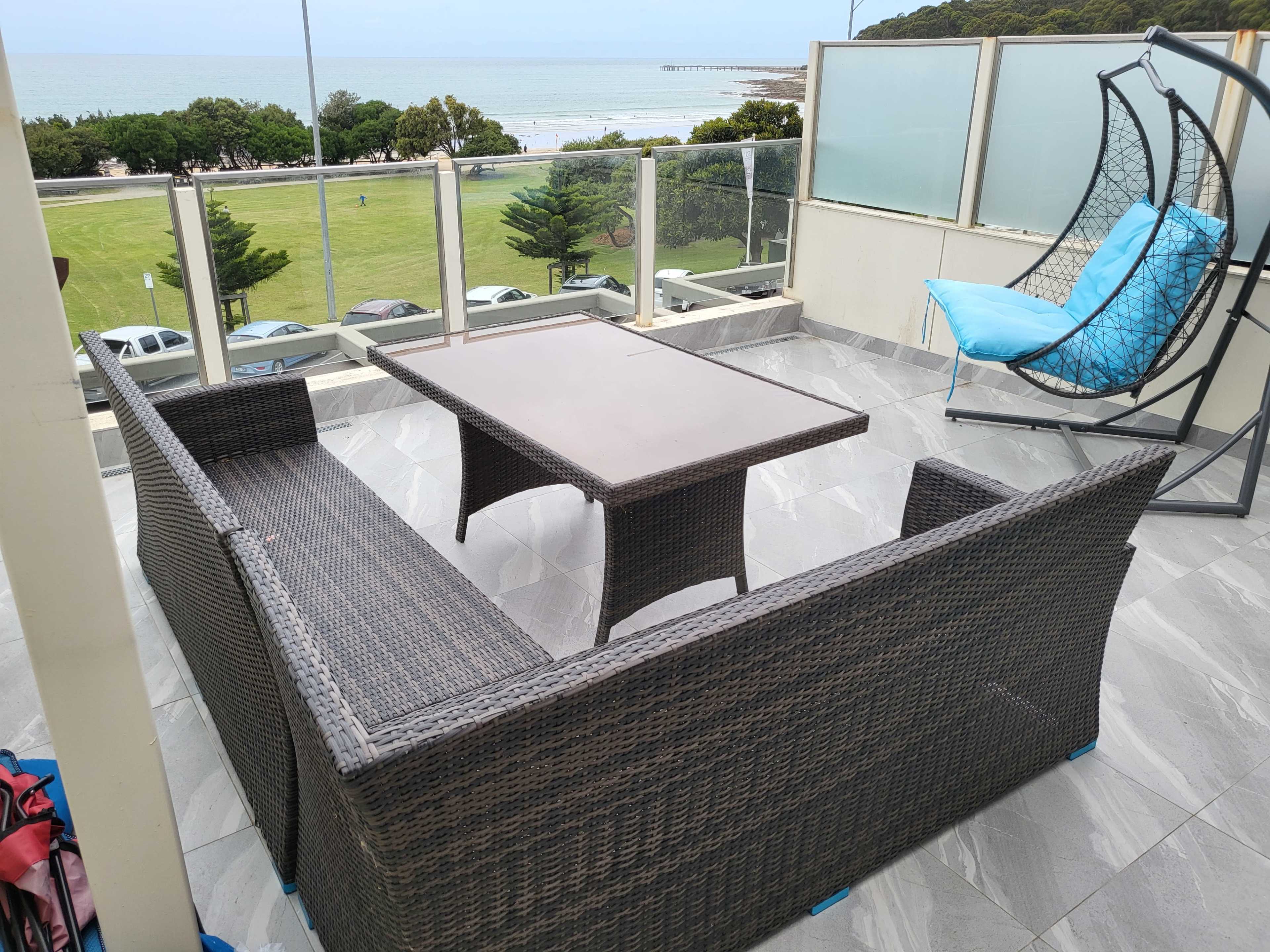 A wicker patio set with a table and two benches overlooks a grassy area and the ocean in the background.