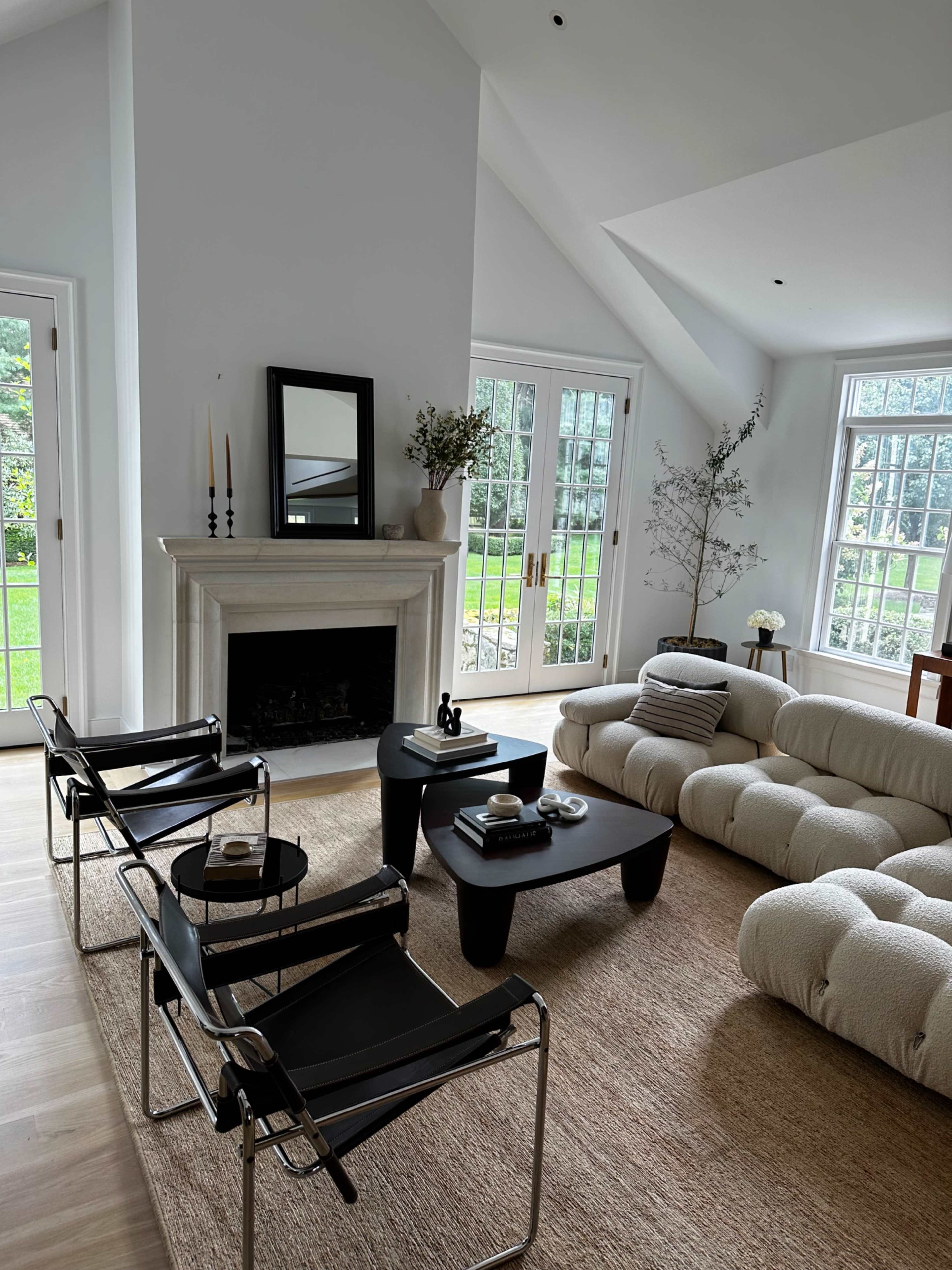 A modern living room with a light-colored couch, black chairs, a coffee table, and large windows overlooking a green garden.