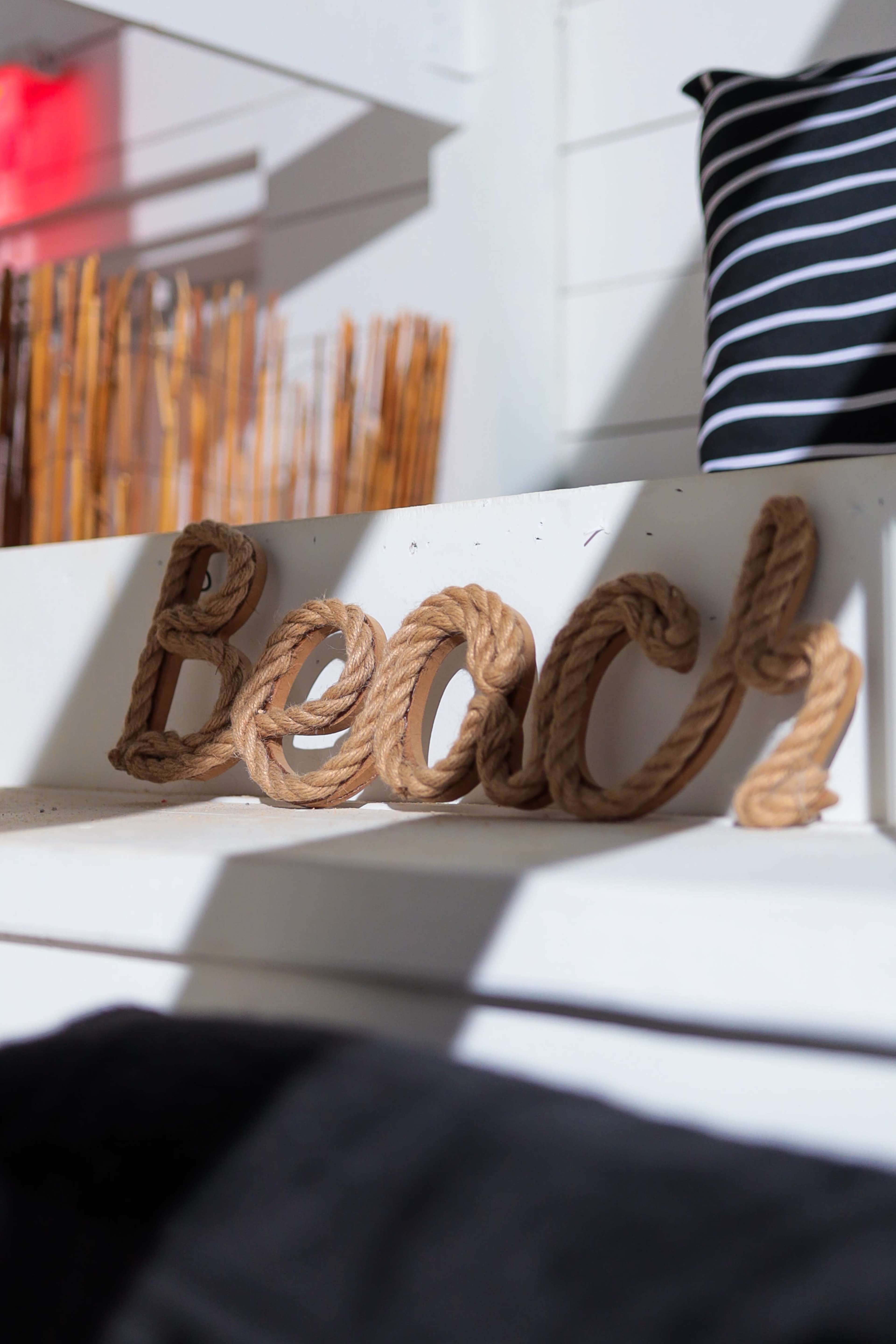 A decorative piece of rope spelling out the word "Beach" is placed on a white shelf, with a striped pillow in the background.