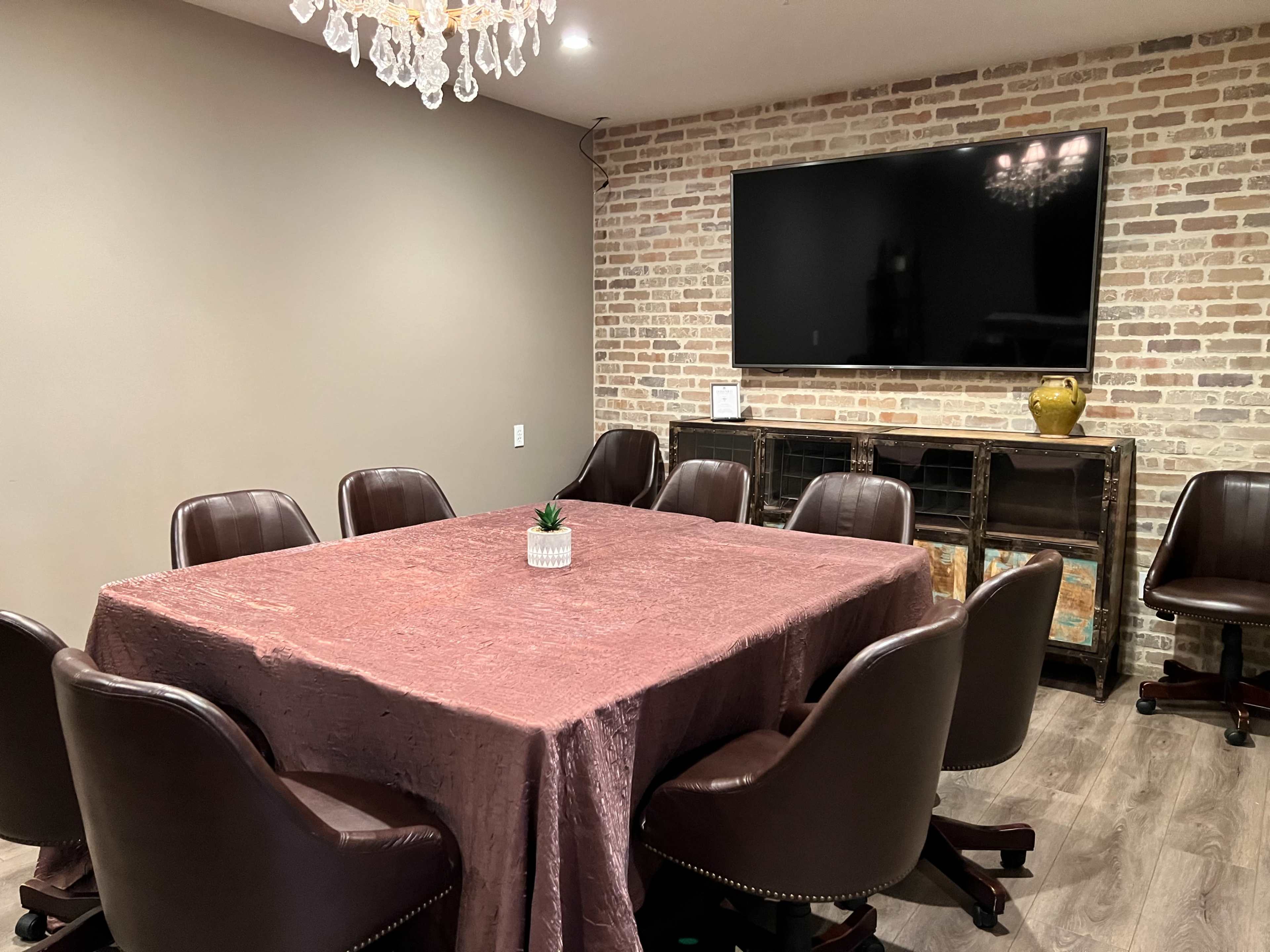 A conference room features a large table surrounded by eight brown chairs, with a brick wall and a television mounted above a side cabinet.