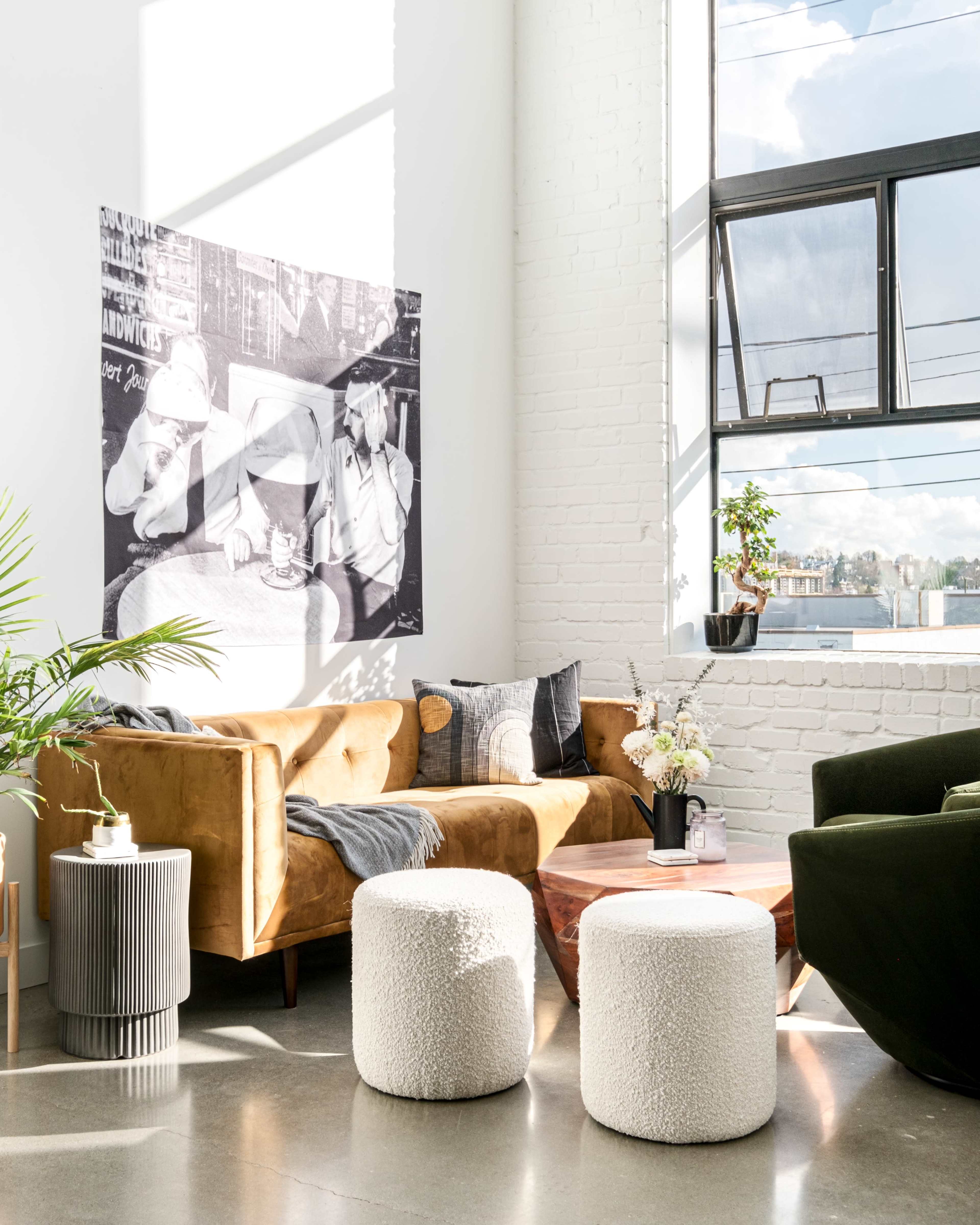 A modern living room features a brown couch, white textured ottomans, and large windows with a black-and-white photograph on the wall.