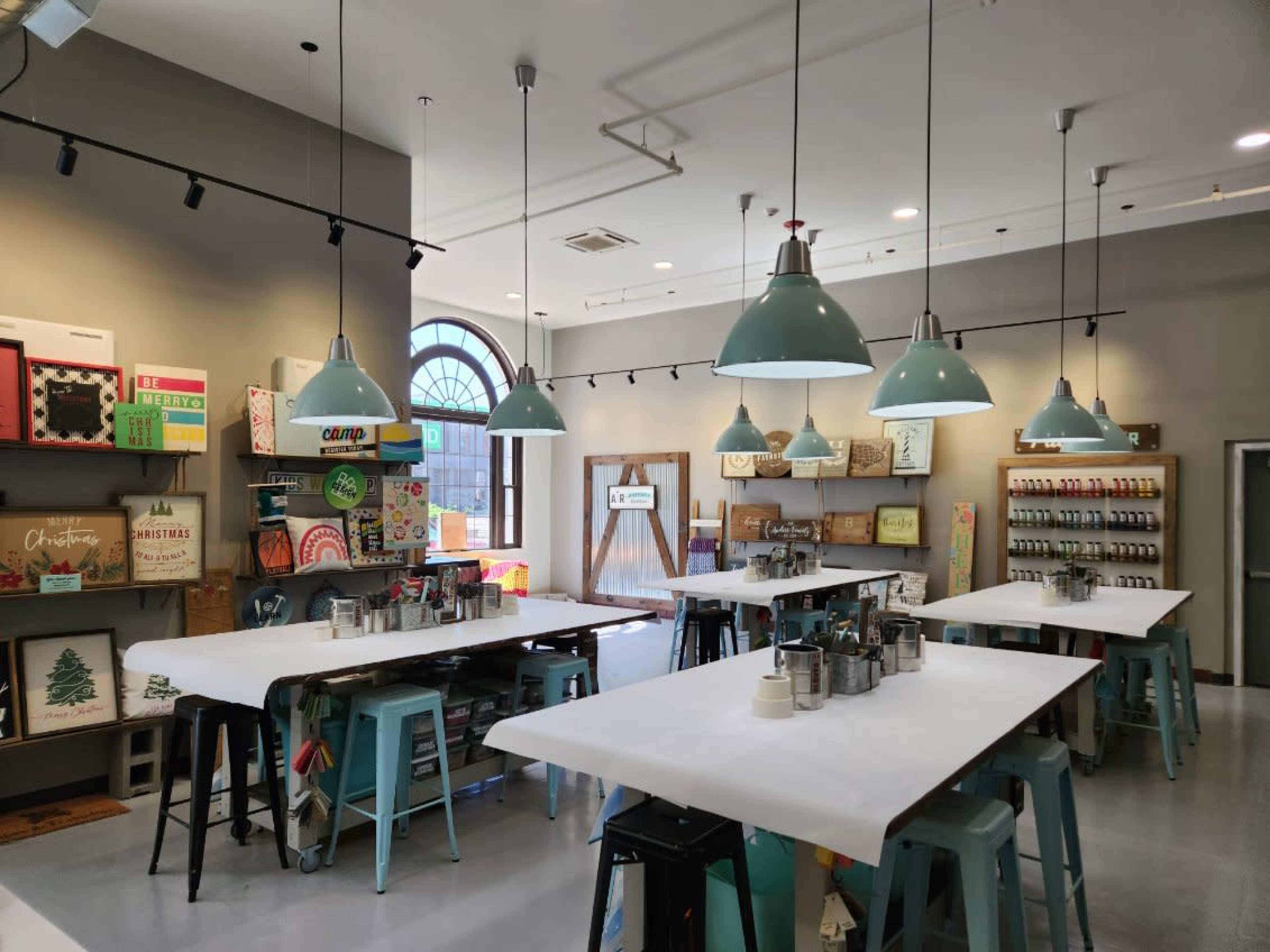 A spacious crafting studio with several tables, teal pendant lights, and shelves displaying various art supplies and decorative items.