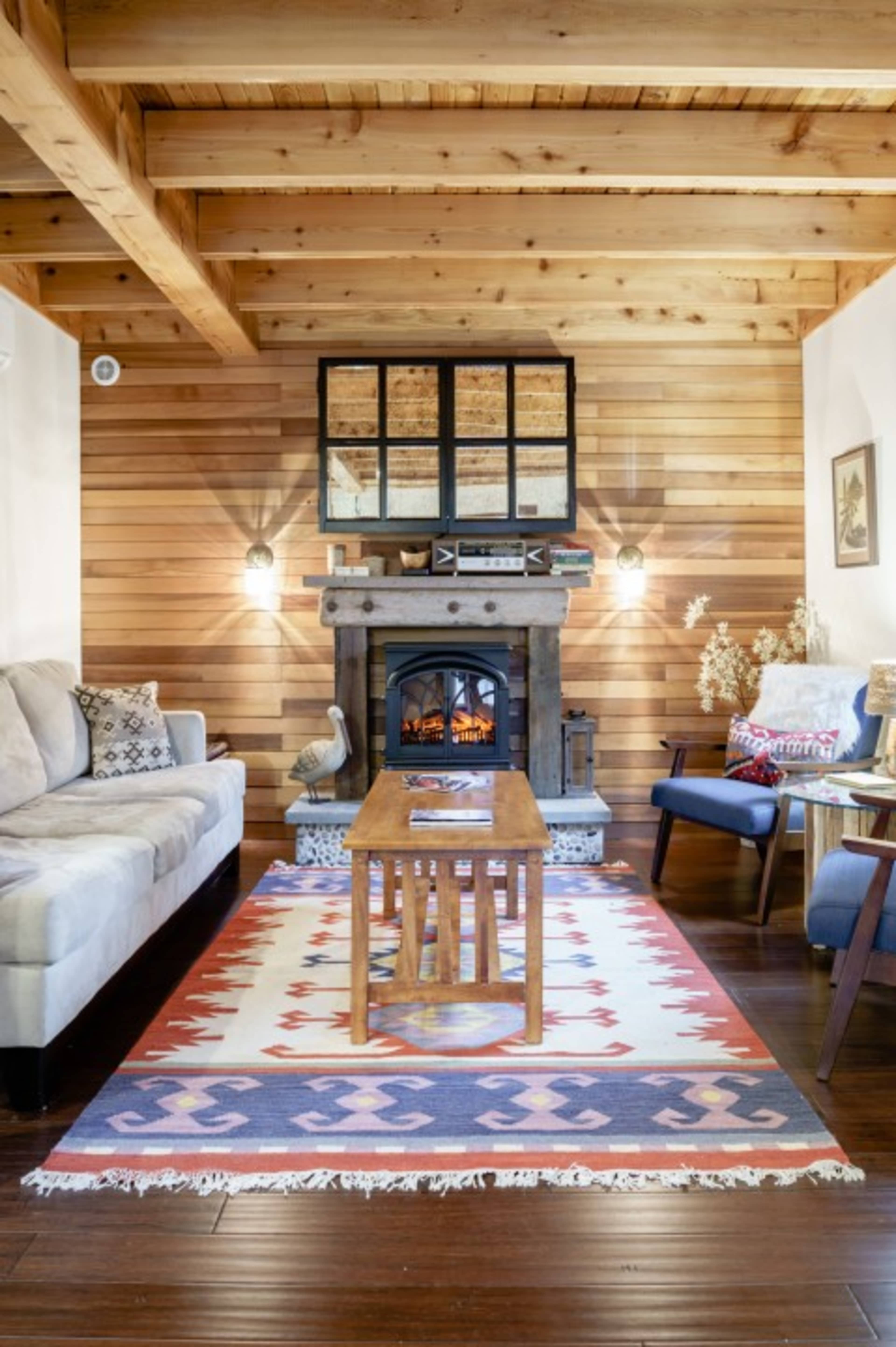 The room features a stone fireplace with a wooden mantel, surrounded by a rustic wooden wall, a couch, a coffee table, and a patterned area rug.