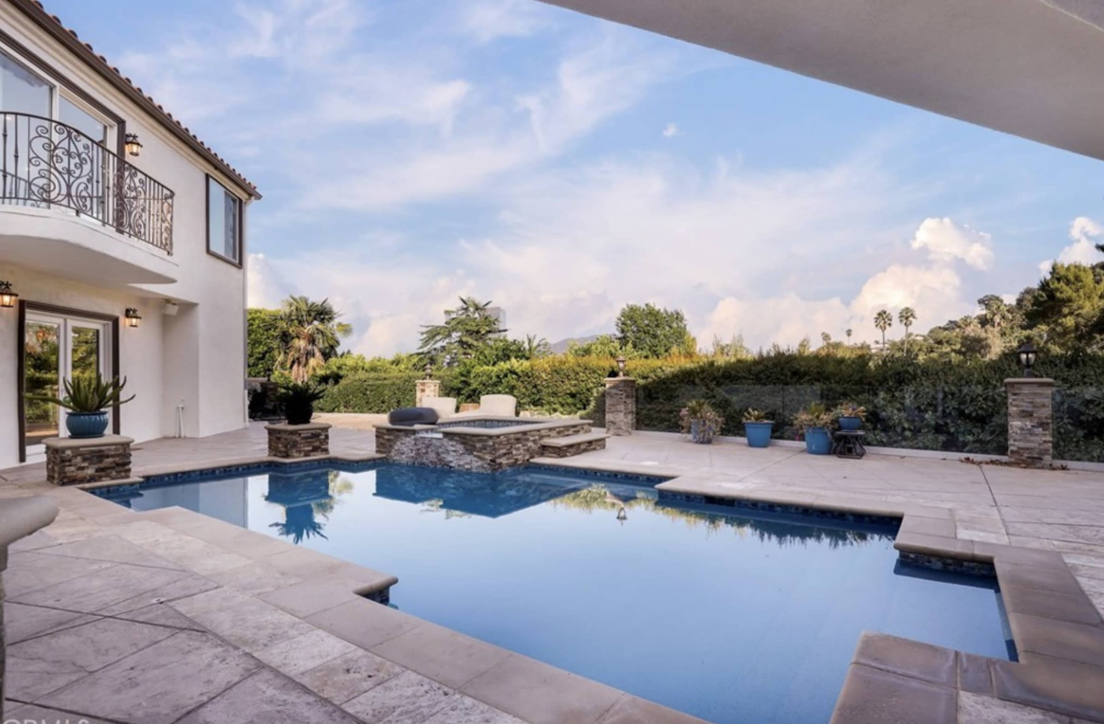 The image shows a modern patio with a swimming pool surrounded by stone features and potted plants, overlooked by a two-story house and a clear sky.