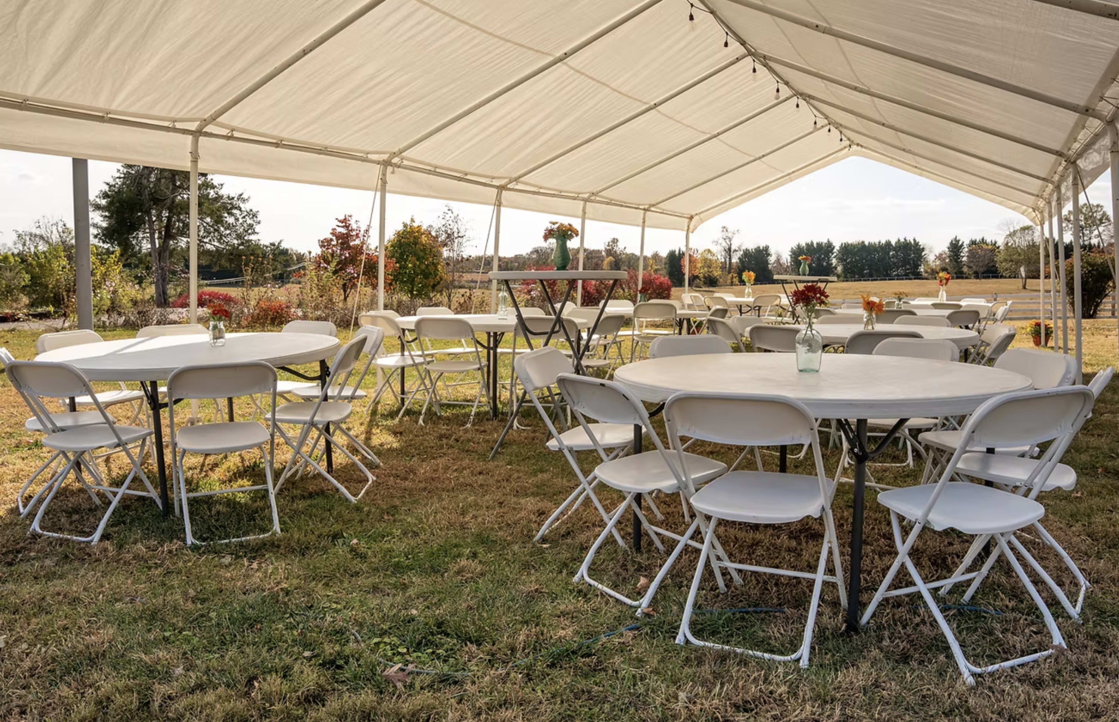 A large white tent is set up outdoors with several round tables and white chairs arranged around them, surrounded by greenery and floral decorations.
