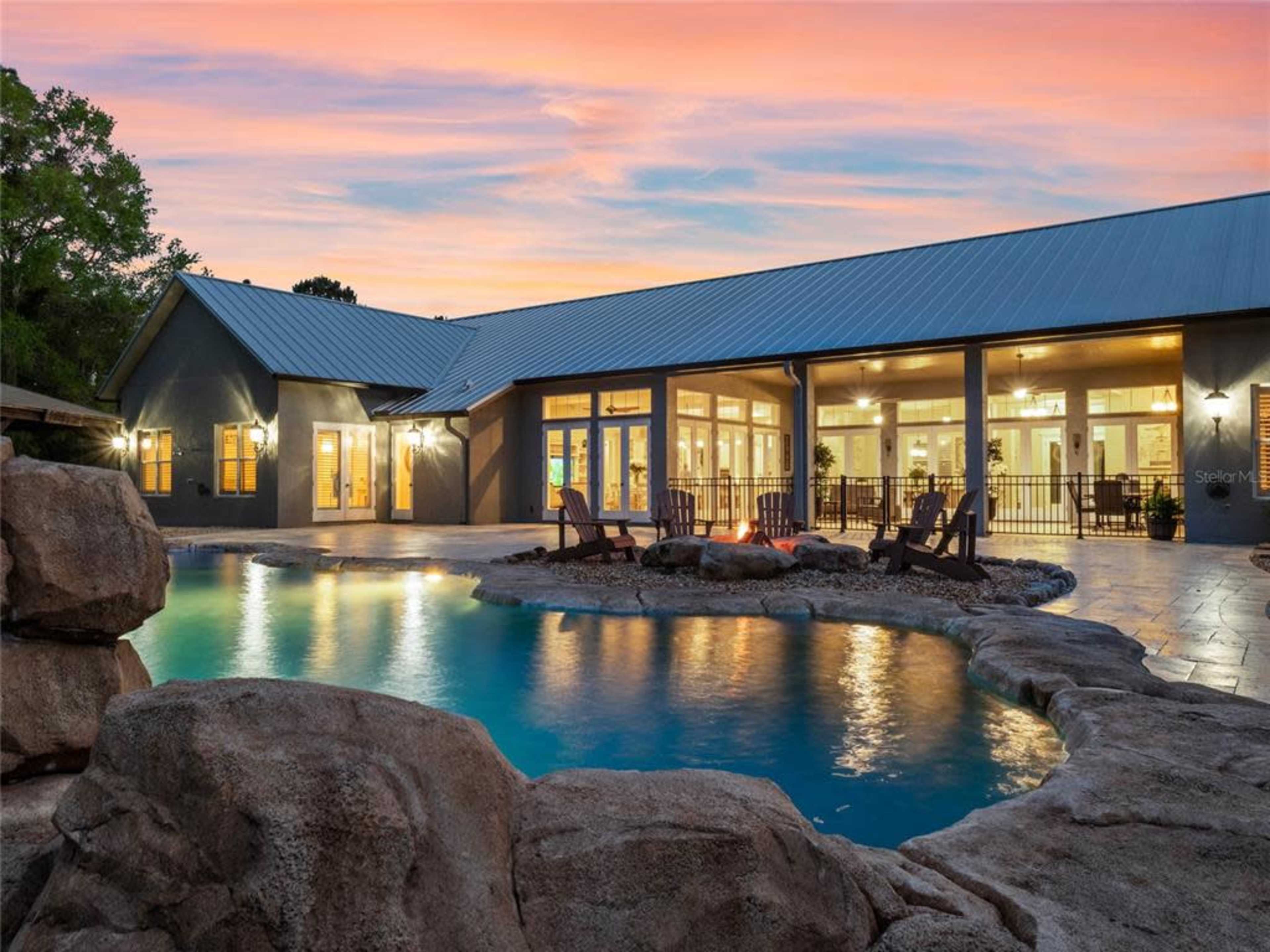 The image shows a modern house with a large patio and swimming pool surrounded by rock formations under a colorful sunset.