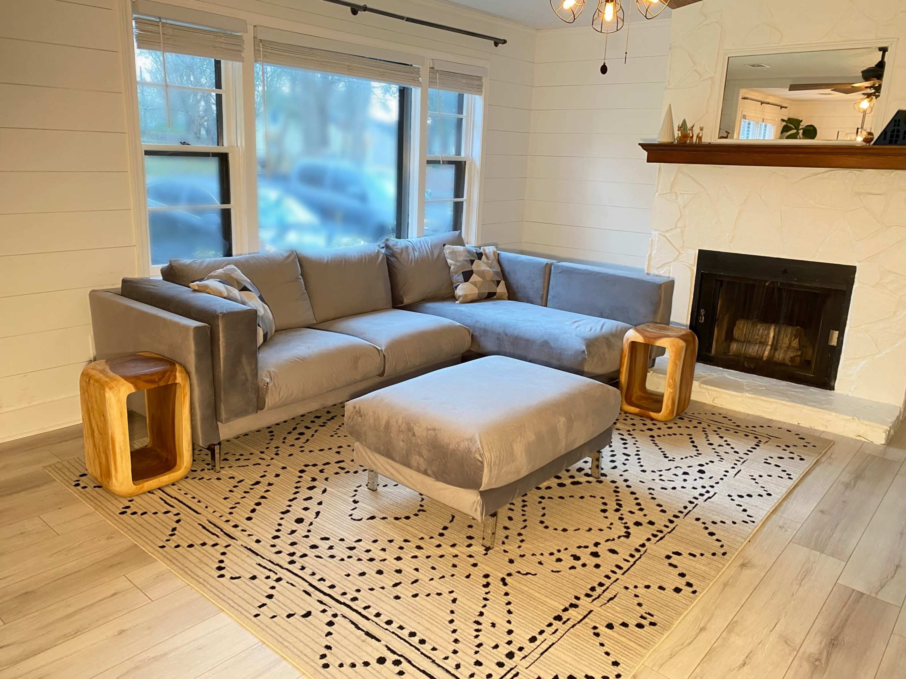 A gray sectional sofa with a matching ottoman is positioned in a bright living room featuring a stone fireplace and wooden accent tables.