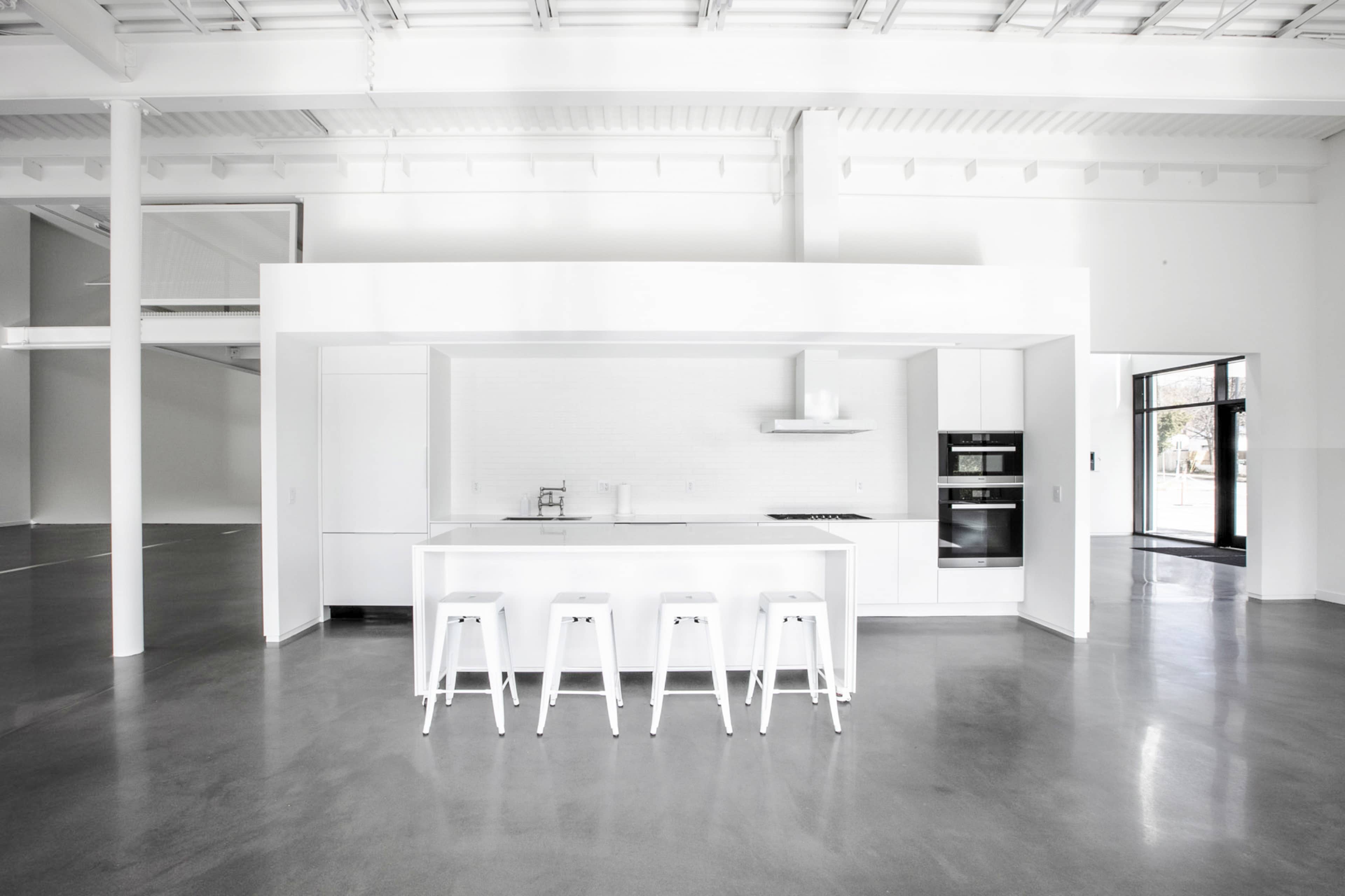 The image depicts a modern kitchen with a minimalist design, featuring a white island and four white stools, set in a spacious, light-filled area with concrete floors.