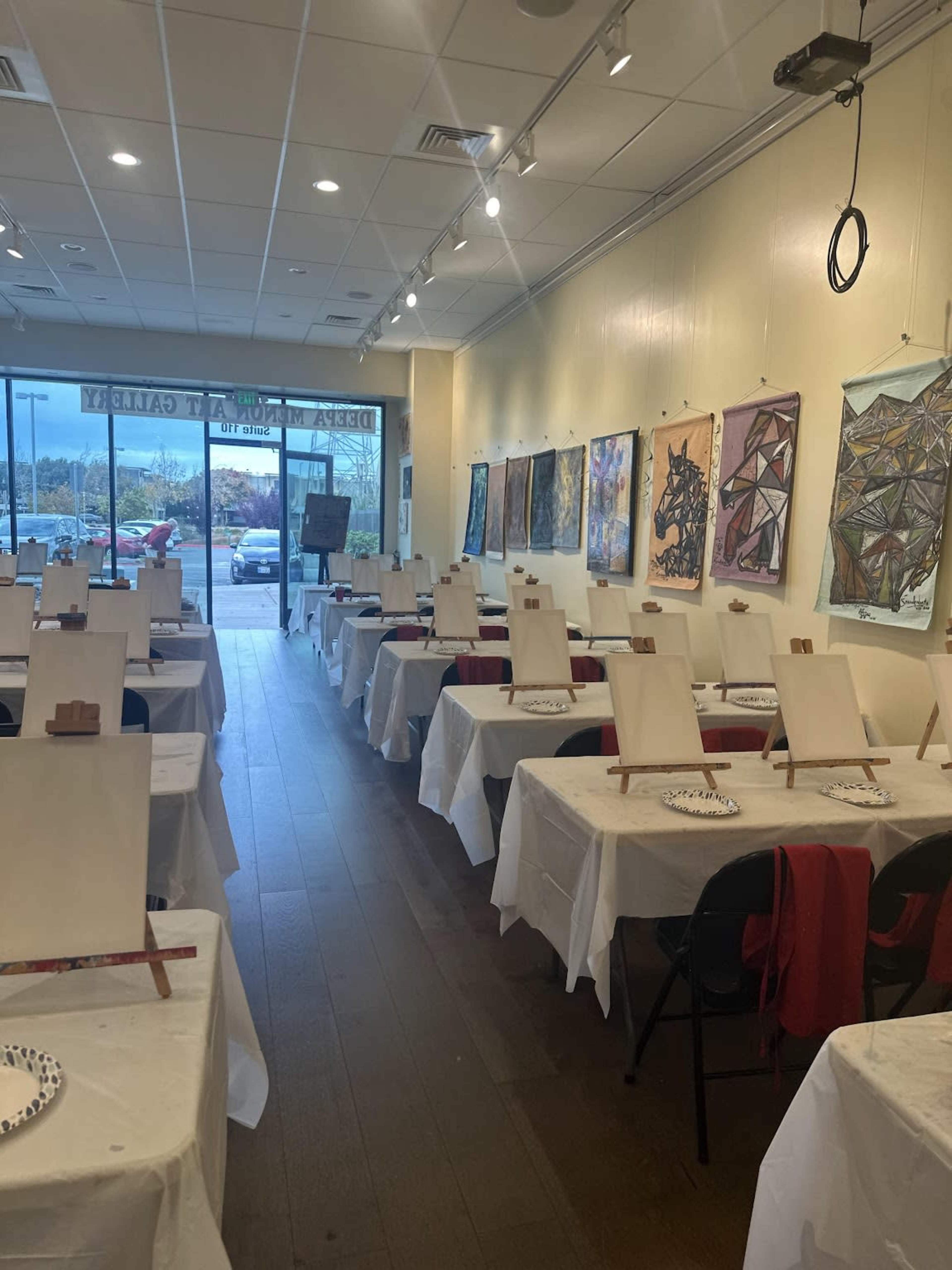 A spacious art studio with tables covered in white cloths, each set up with blank canvases and palettes, surrounded by colorful artwork displayed on the walls.