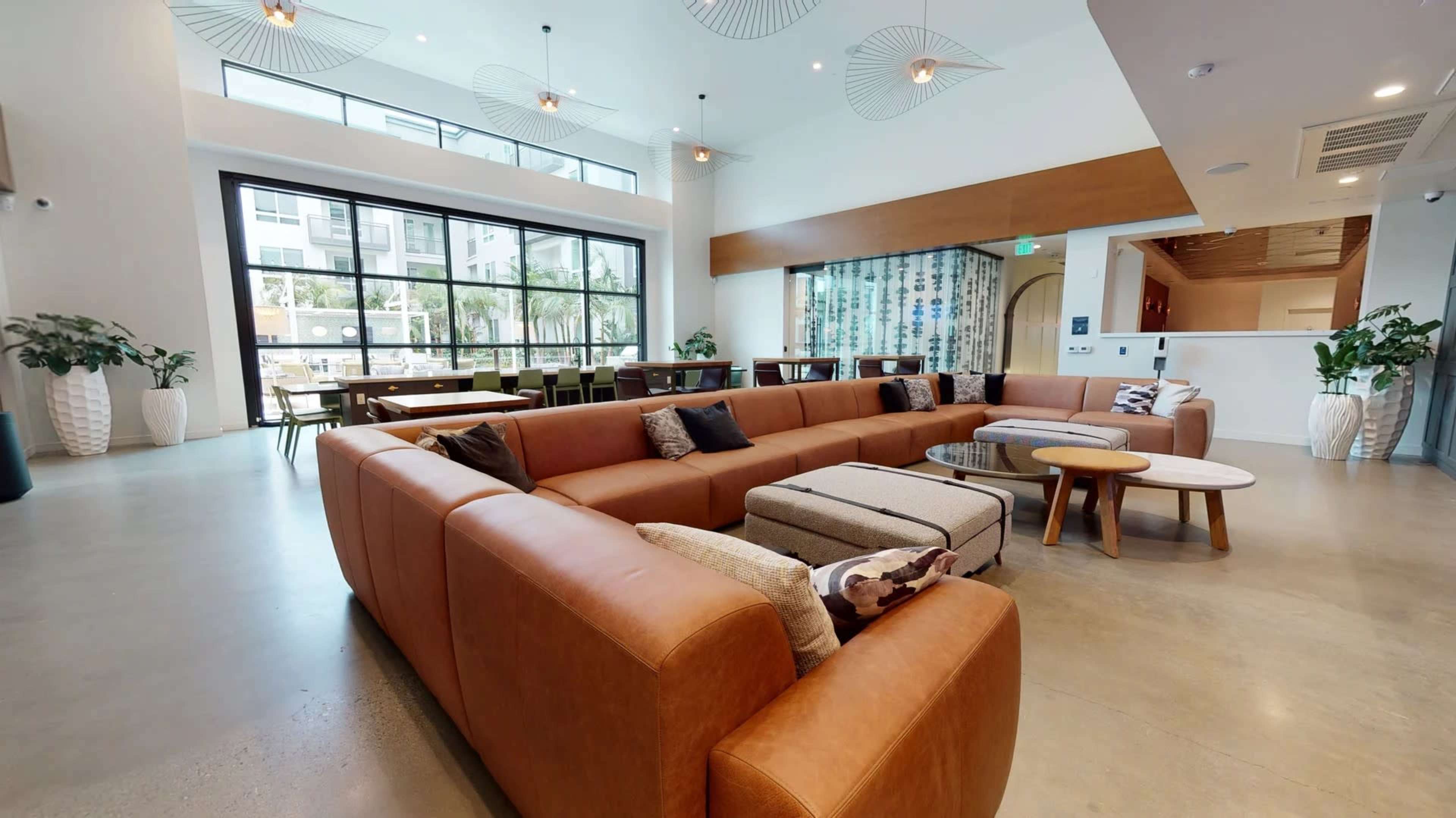 A spacious lounge area features a large, sectional leather sofa, several round tables, and plants, with large windows allowing natural light to illuminate the space.