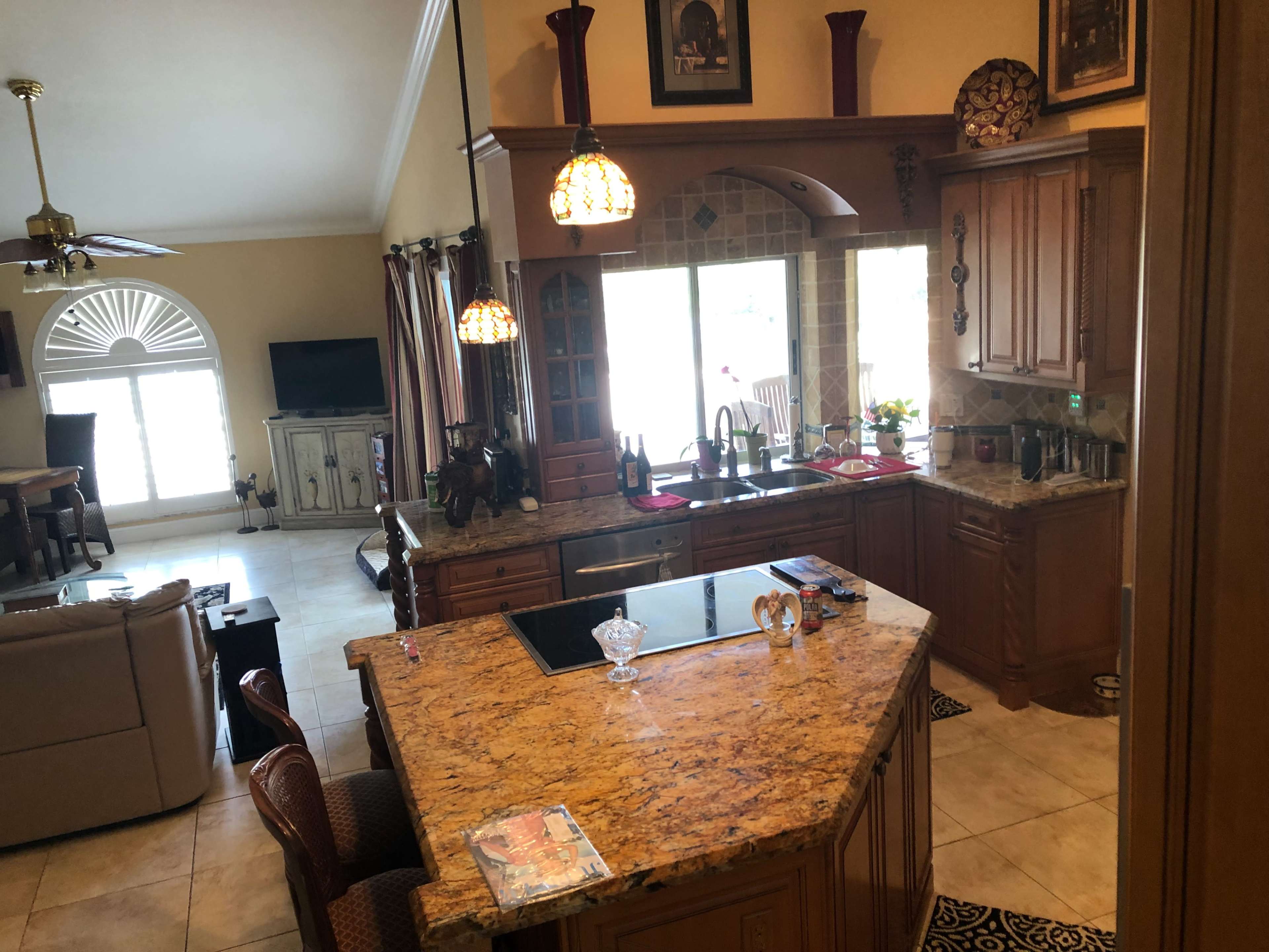 A spacious kitchen features a central island with granite countertops, wooden cabinetry, and large windows overlooking an adjoining living area.