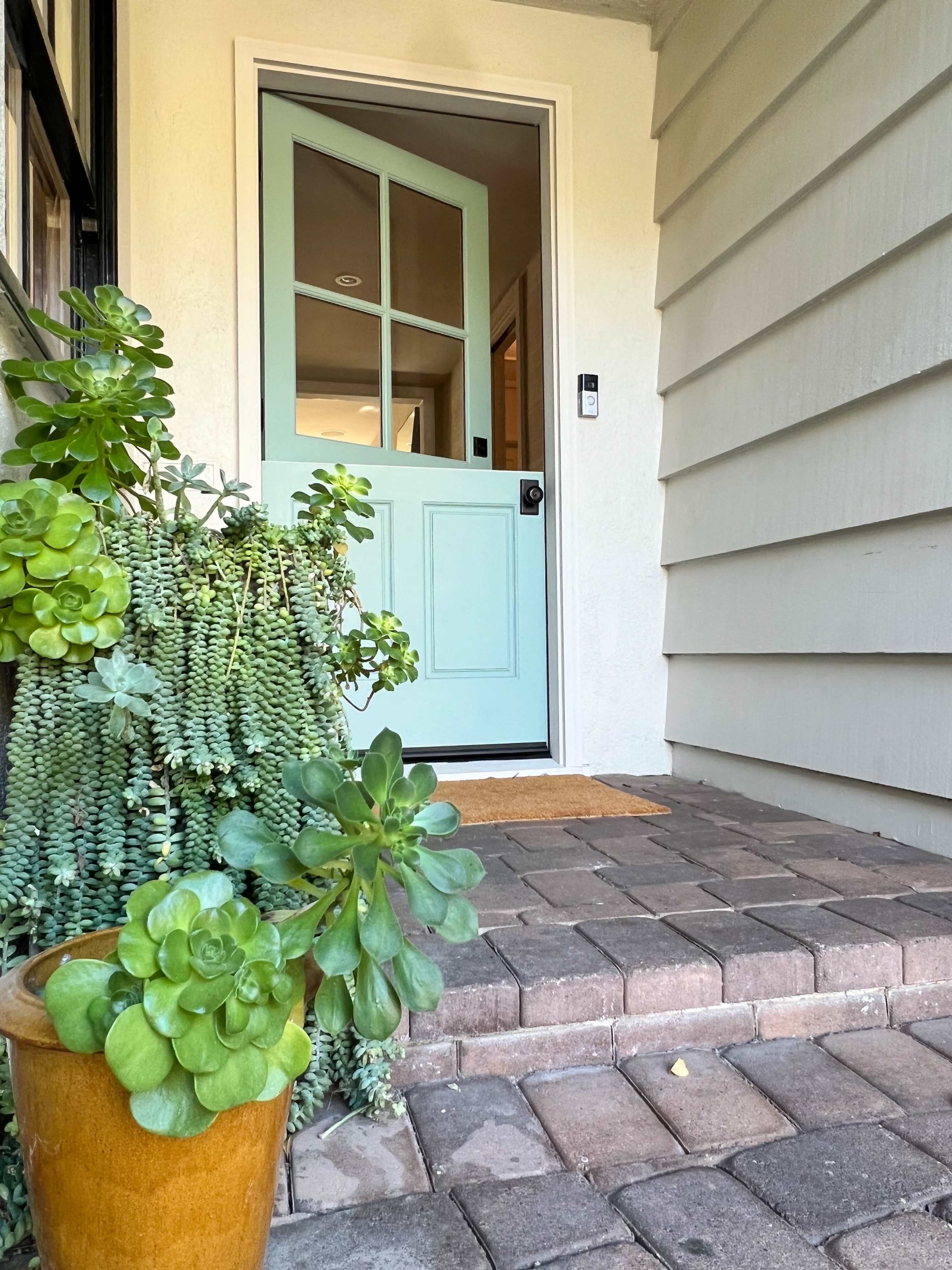 A mint green front door opens to a well-maintained entrance surrounded by potted plants and brick step paths.