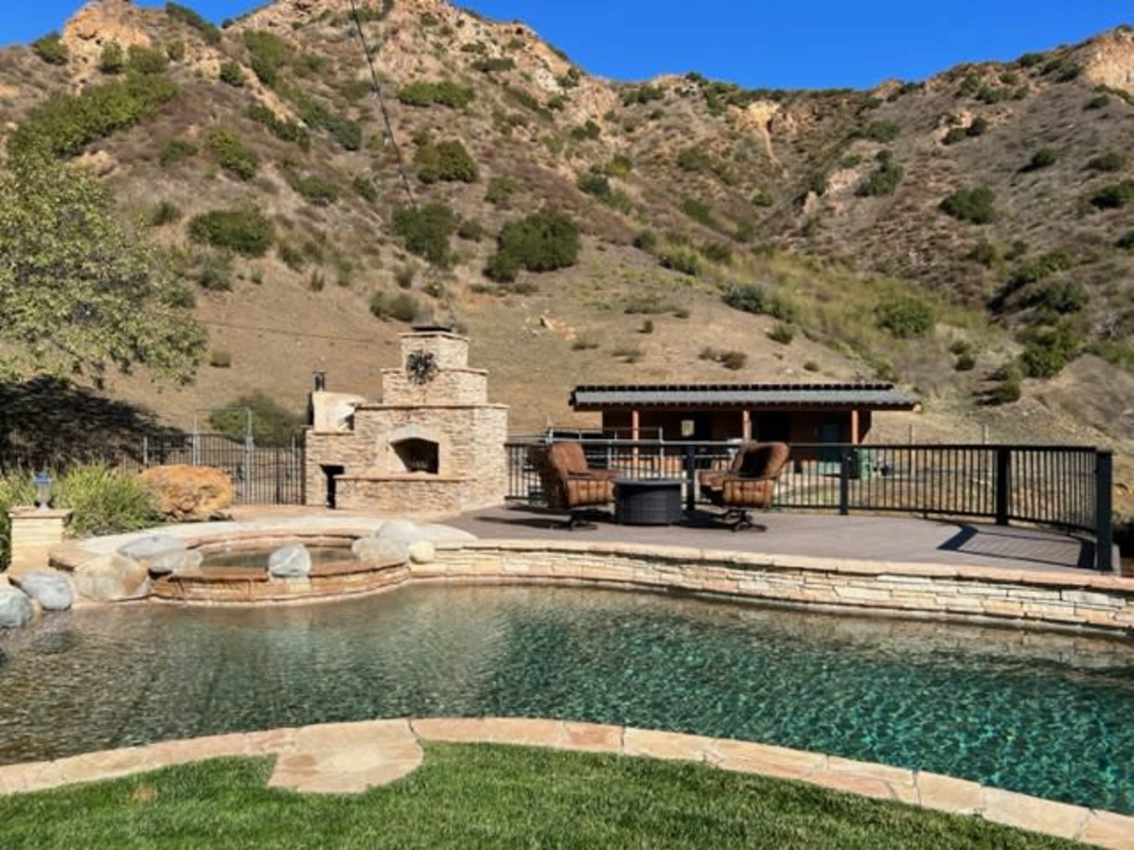 A landscaped backyard with a pool, stone fireplace, seating area, and a backdrop of hills.