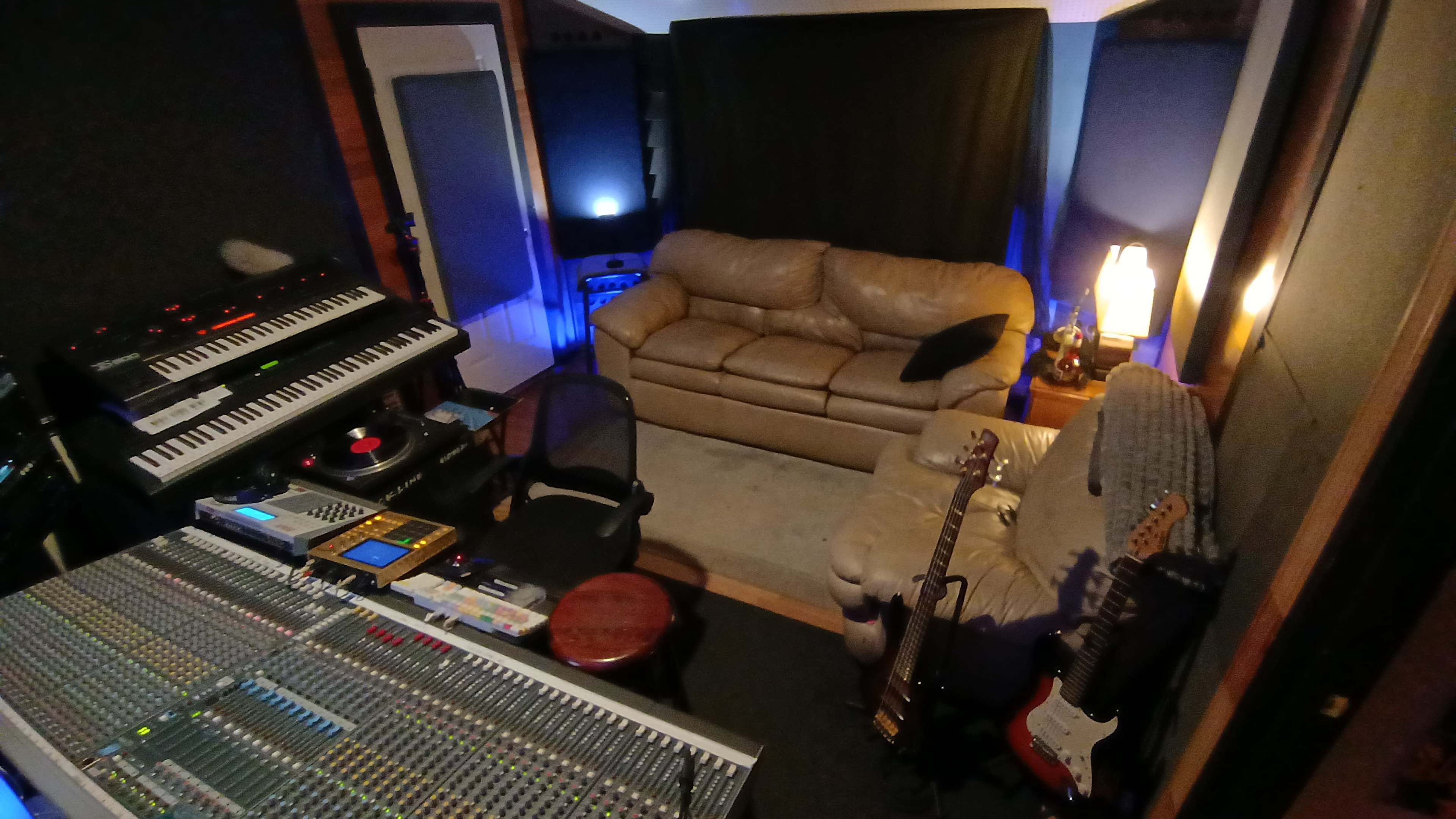The image shows a music recording studio featuring a mixing console, a couch, and several musical instruments.
