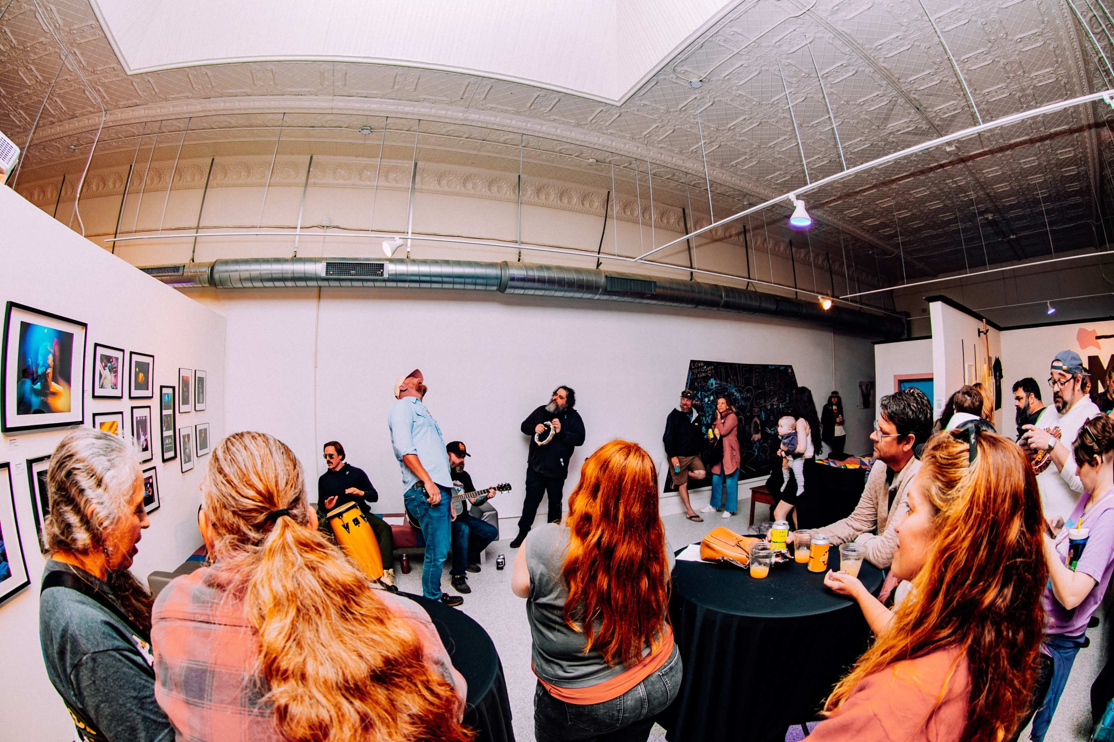 A group of people gathers in an art gallery for an event featuring live music and displayed photographs.
