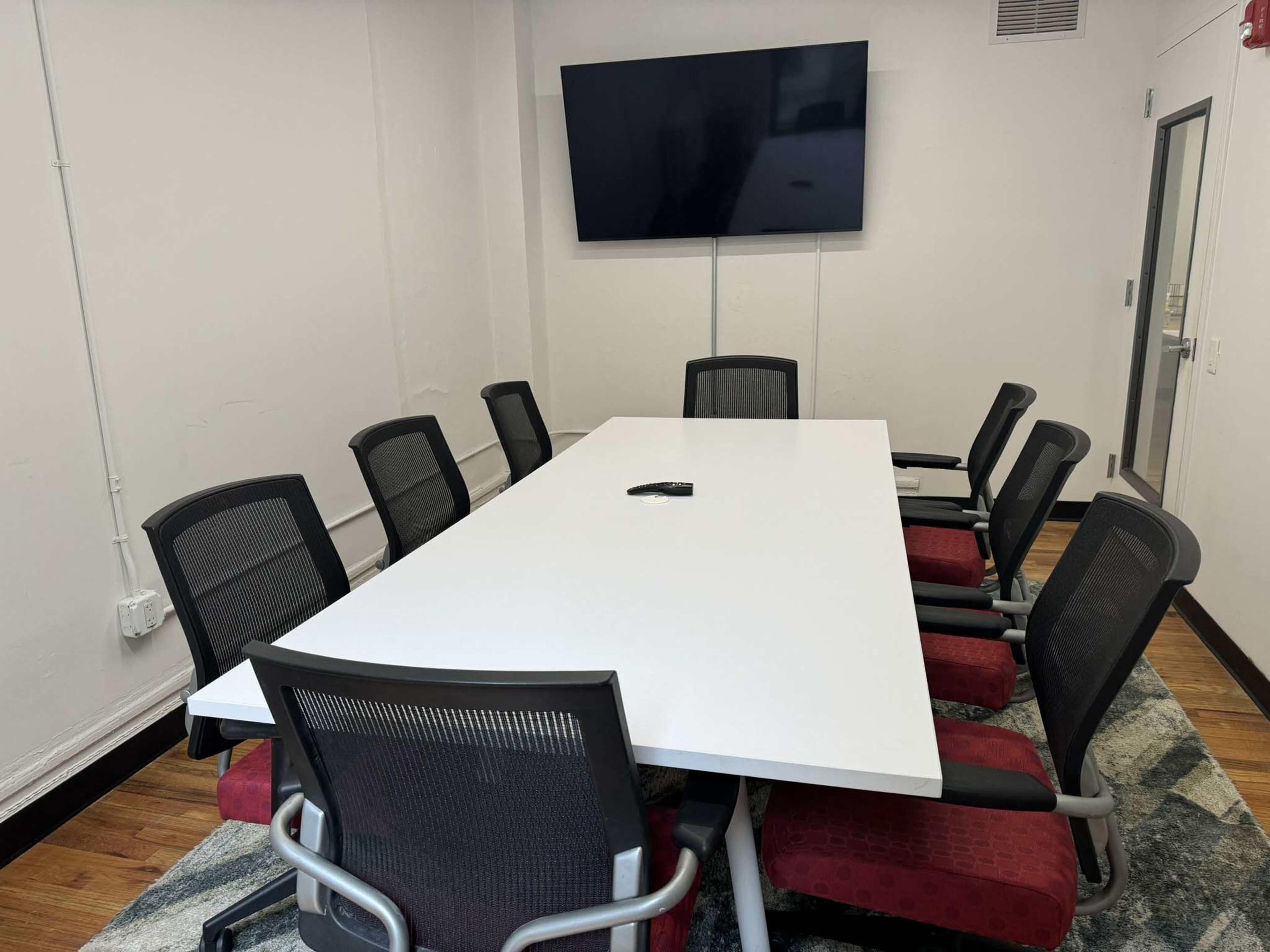 A large white conference table surrounded by eight black mesh chairs is positioned in a brightly lit meeting room with a wall-mounted television.