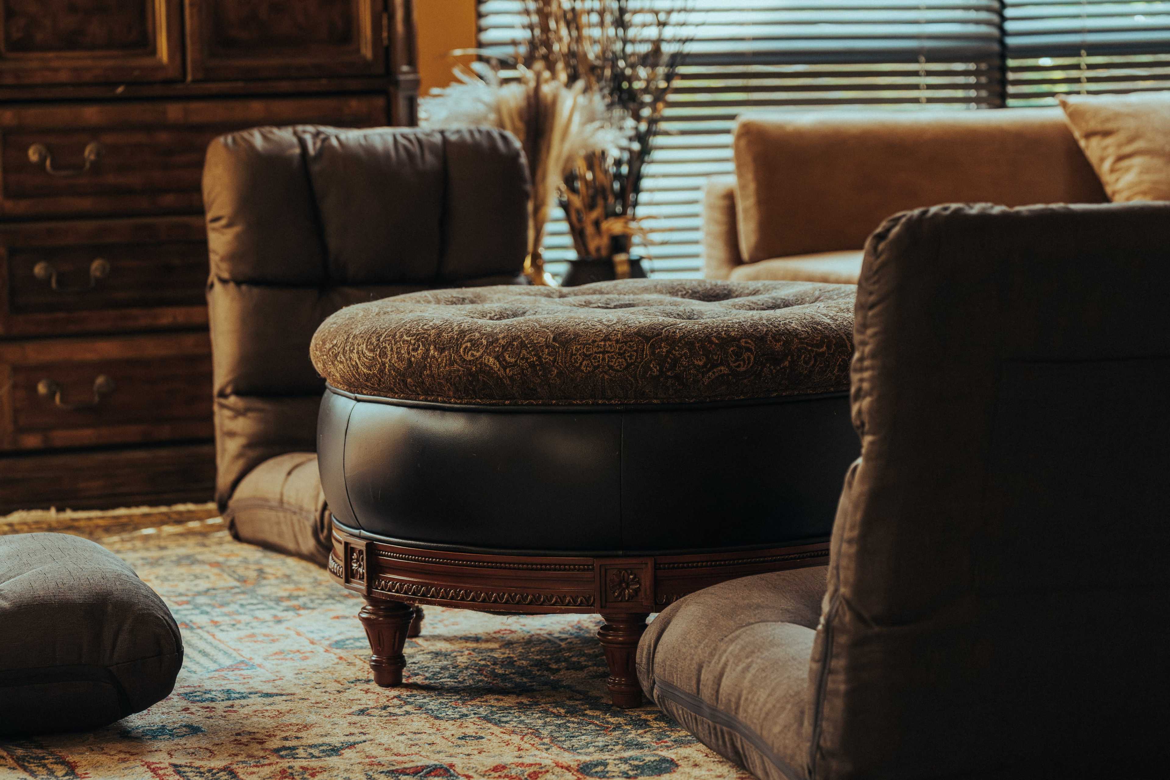 The image depicts a cozy living room with a round, upholstered ottoman in the center, surrounded by low cushioned chairs and various decorative elements.