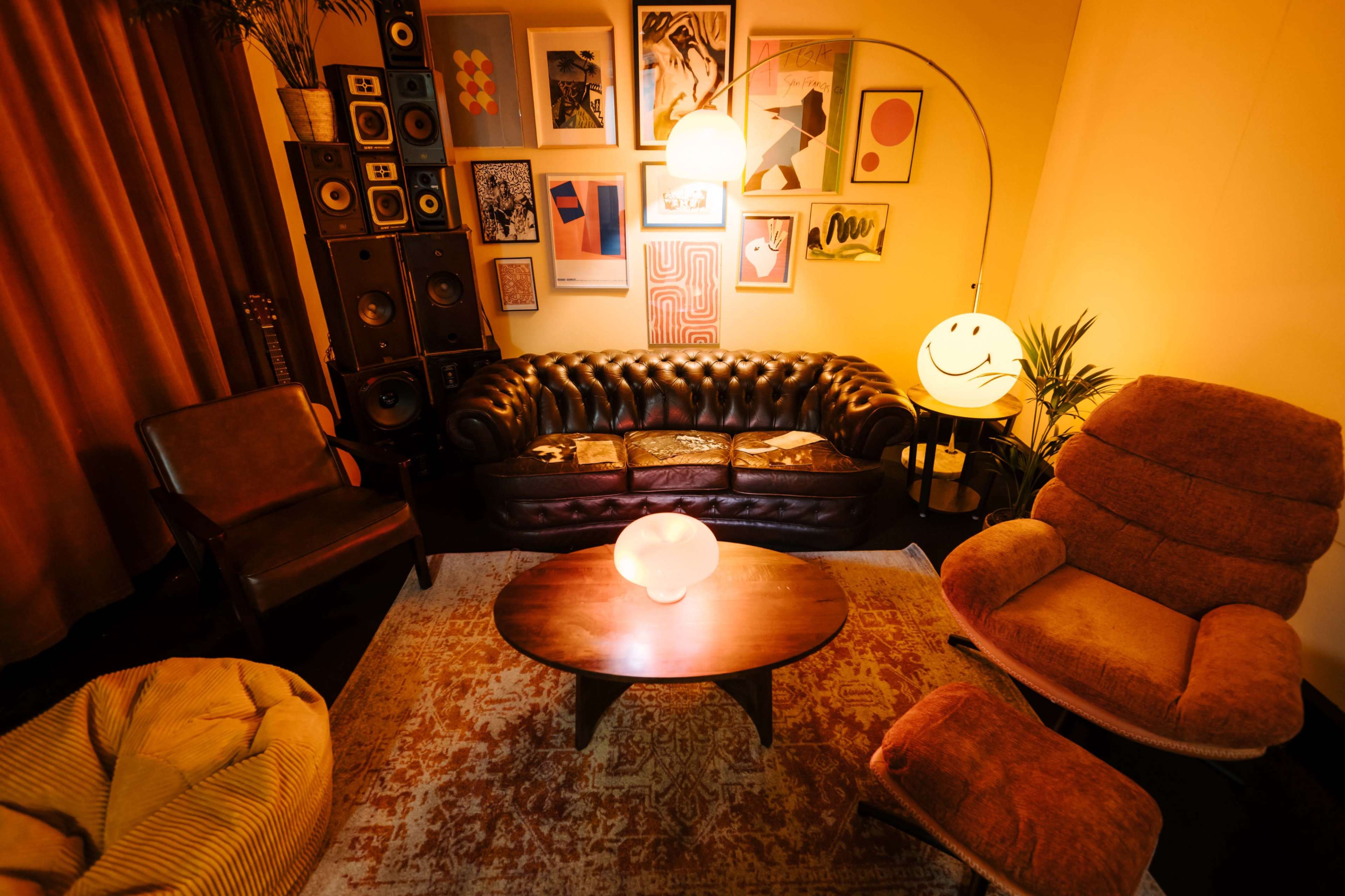 The image features a cozy living room setting with a brown leather couch, a round coffee table, vintage speakers, and warm lighting, adorned with colorful wall art.