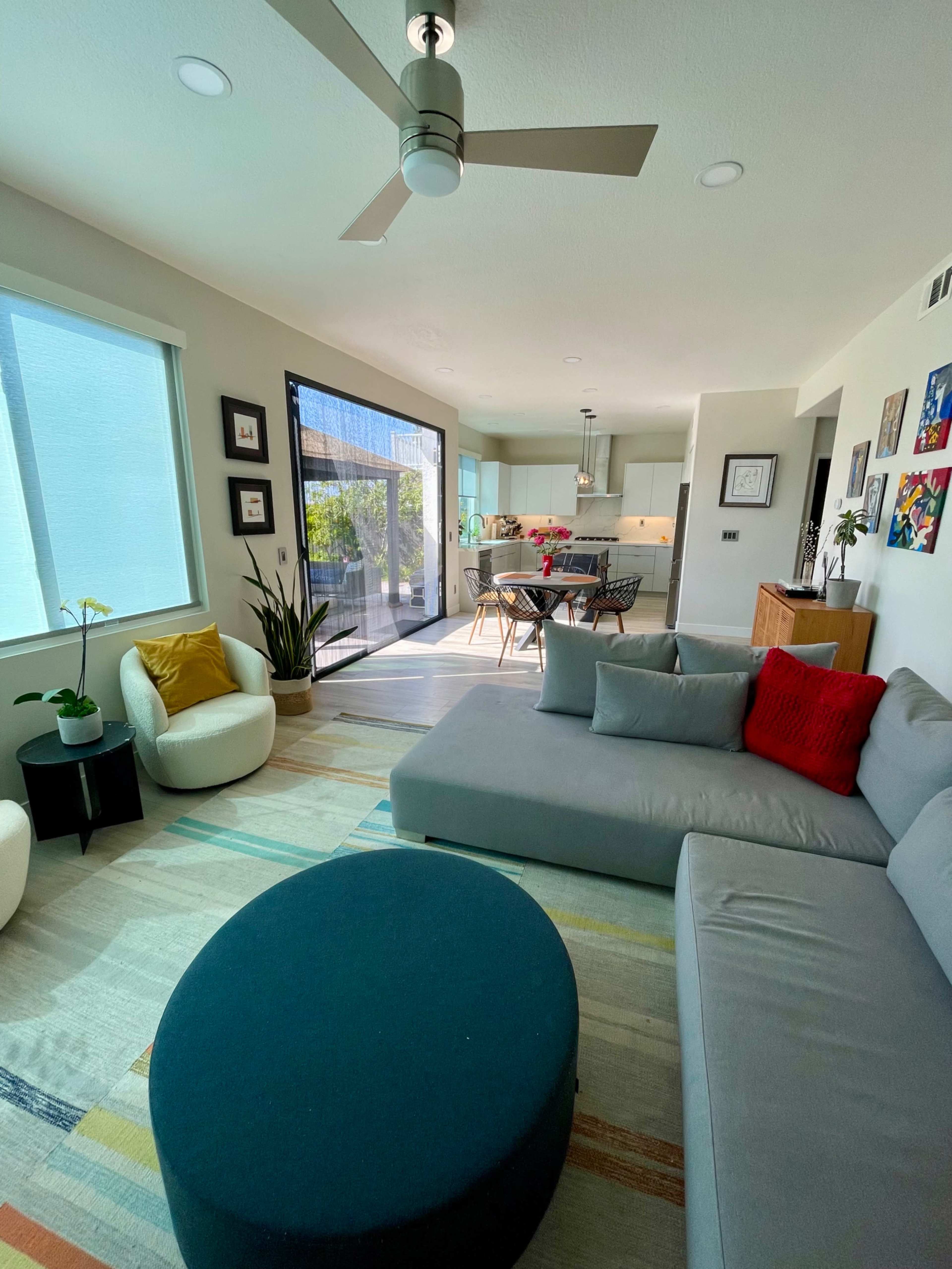 A modern living room with a sectional sofa, an ottoman, and a dining area in an open-concept space, featuring large windows that provide natural light.
