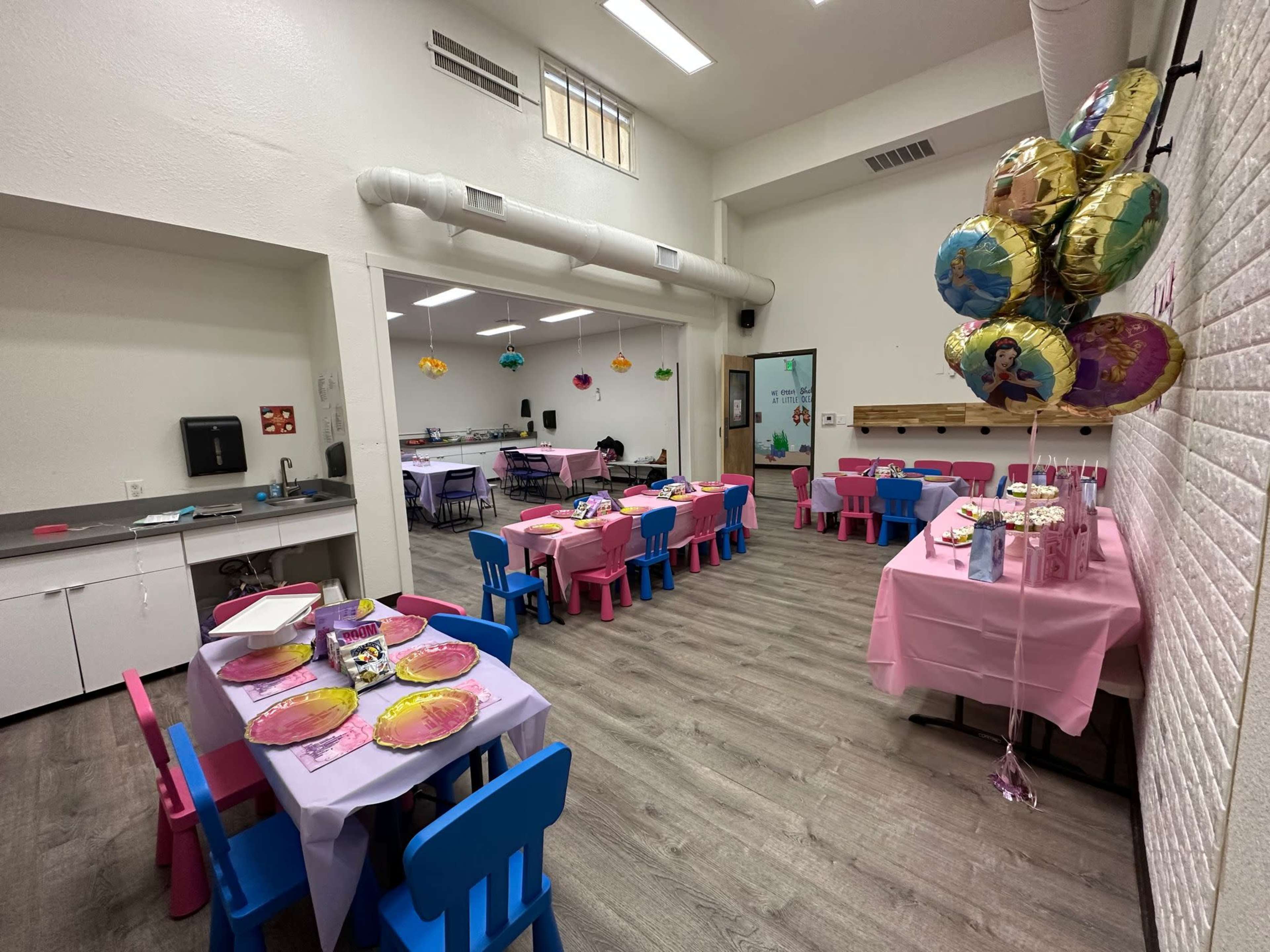 A spacious room is set up with multiple tables covered in pink tablecloths and decorated with colorful balloons.