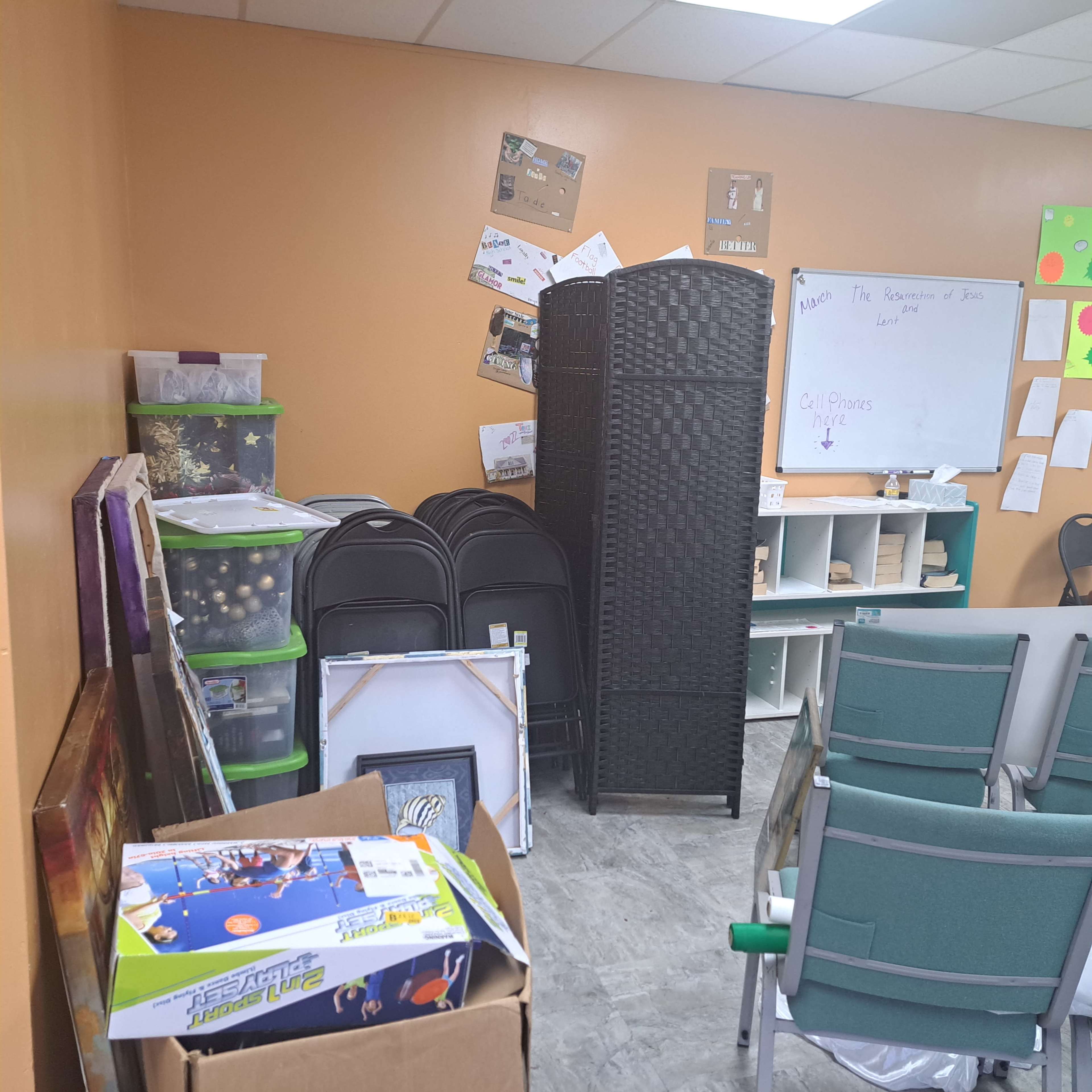 The image shows a cluttered room with stacked folding chairs, storage containers, a room divider, and various items on the walls.