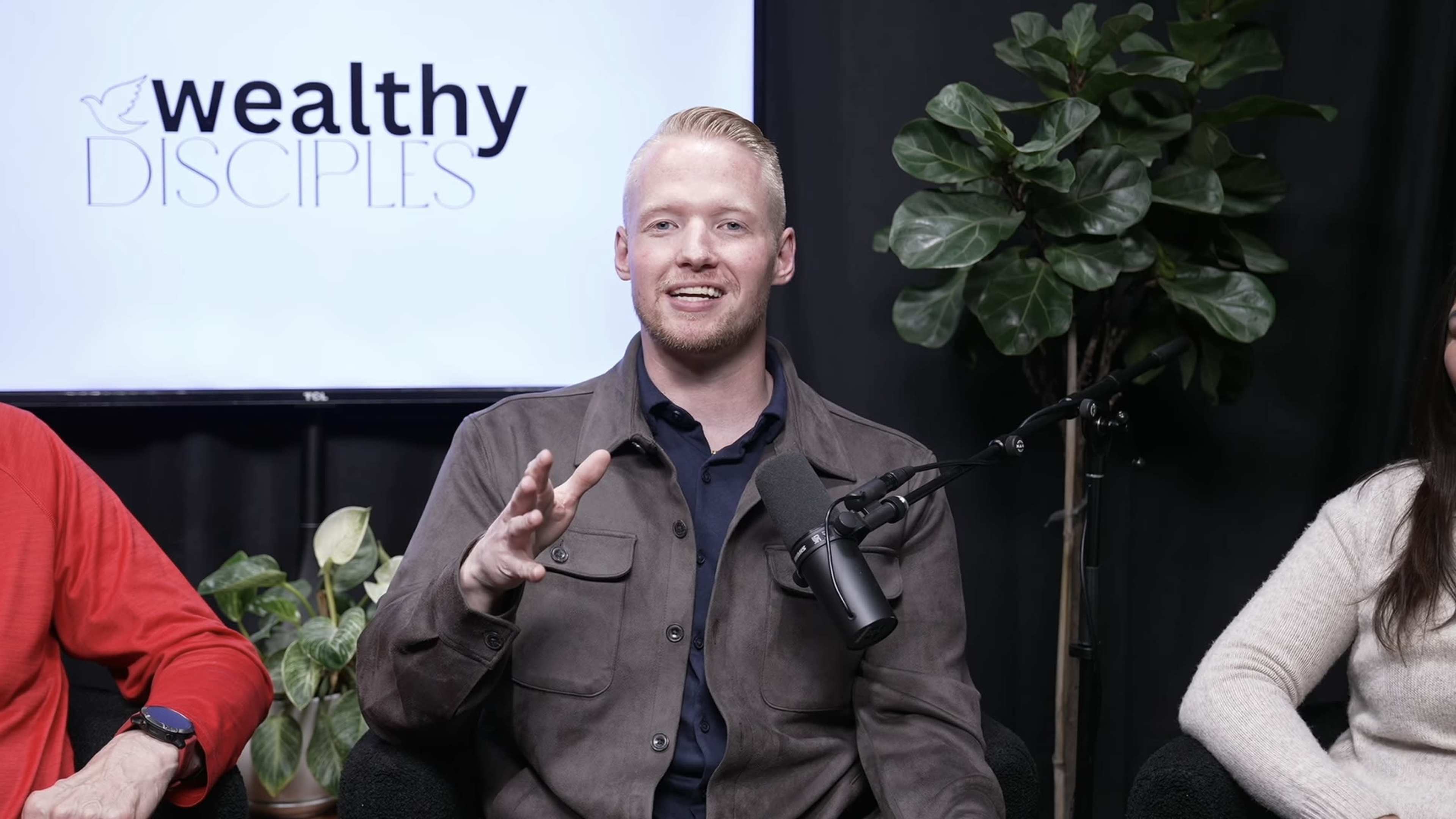 A man is sitting in front of a microphone, speaking while a logo for "Wealthy Disciples" is displayed on a screen behind him, with indoor plants nearby.