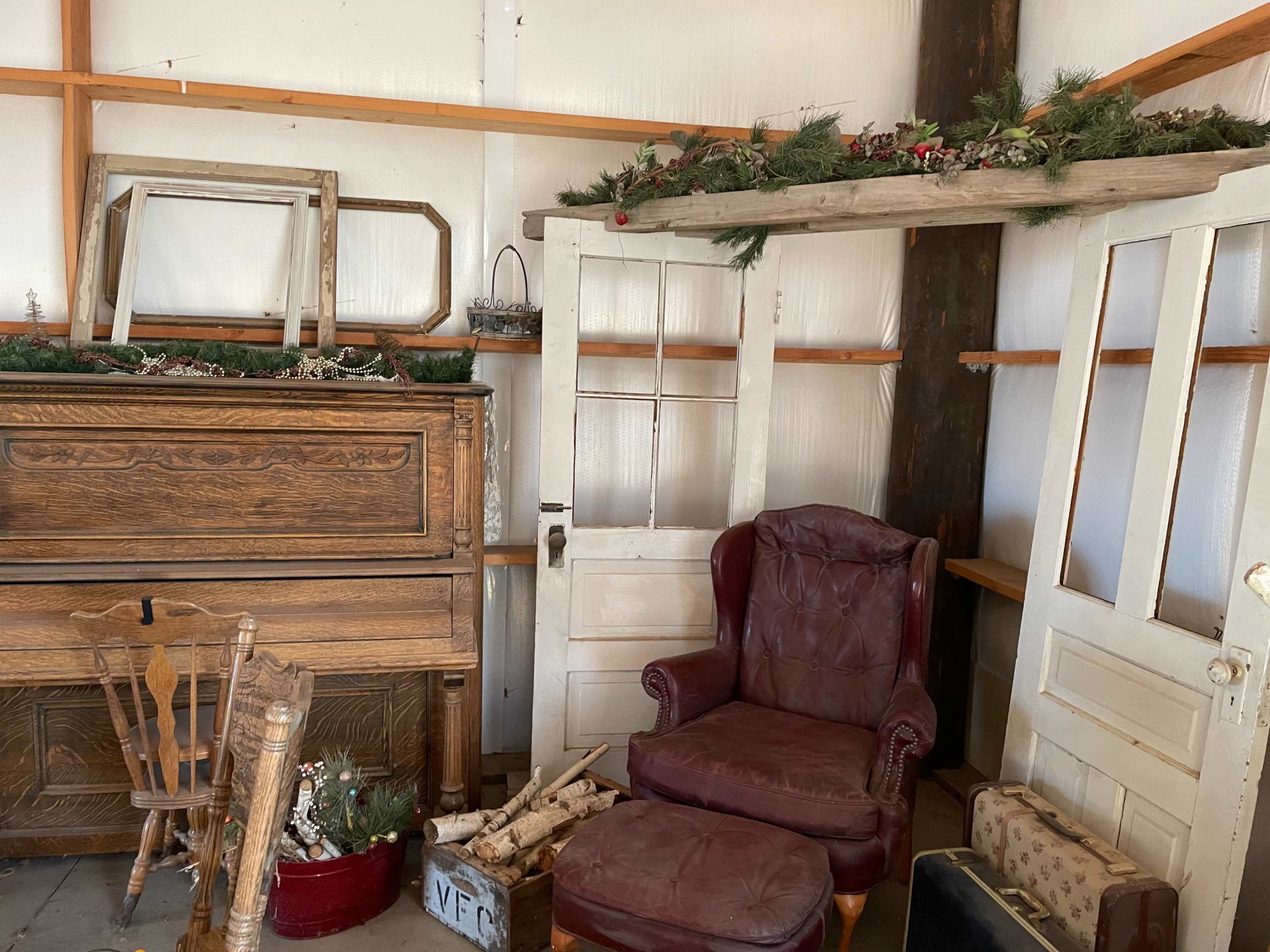 An interior of a storage space with wooden shelves, an old piano, a red leather armchair, a wooden chair, and various decor items, including logs and garlands.