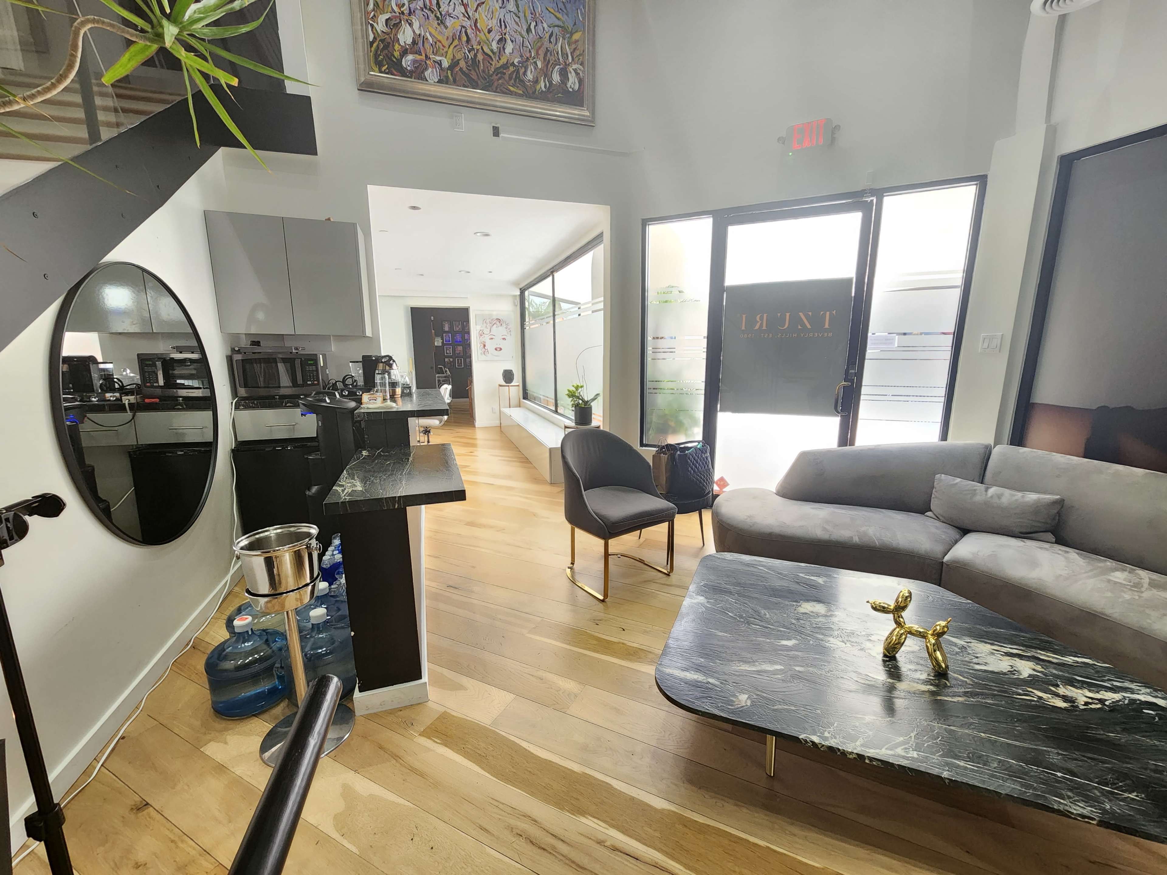 The image shows a contemporary interior space featuring a lounge area with a gray sofa and a black marble coffee table, alongside a kitchenette area and large windows for natural light.
