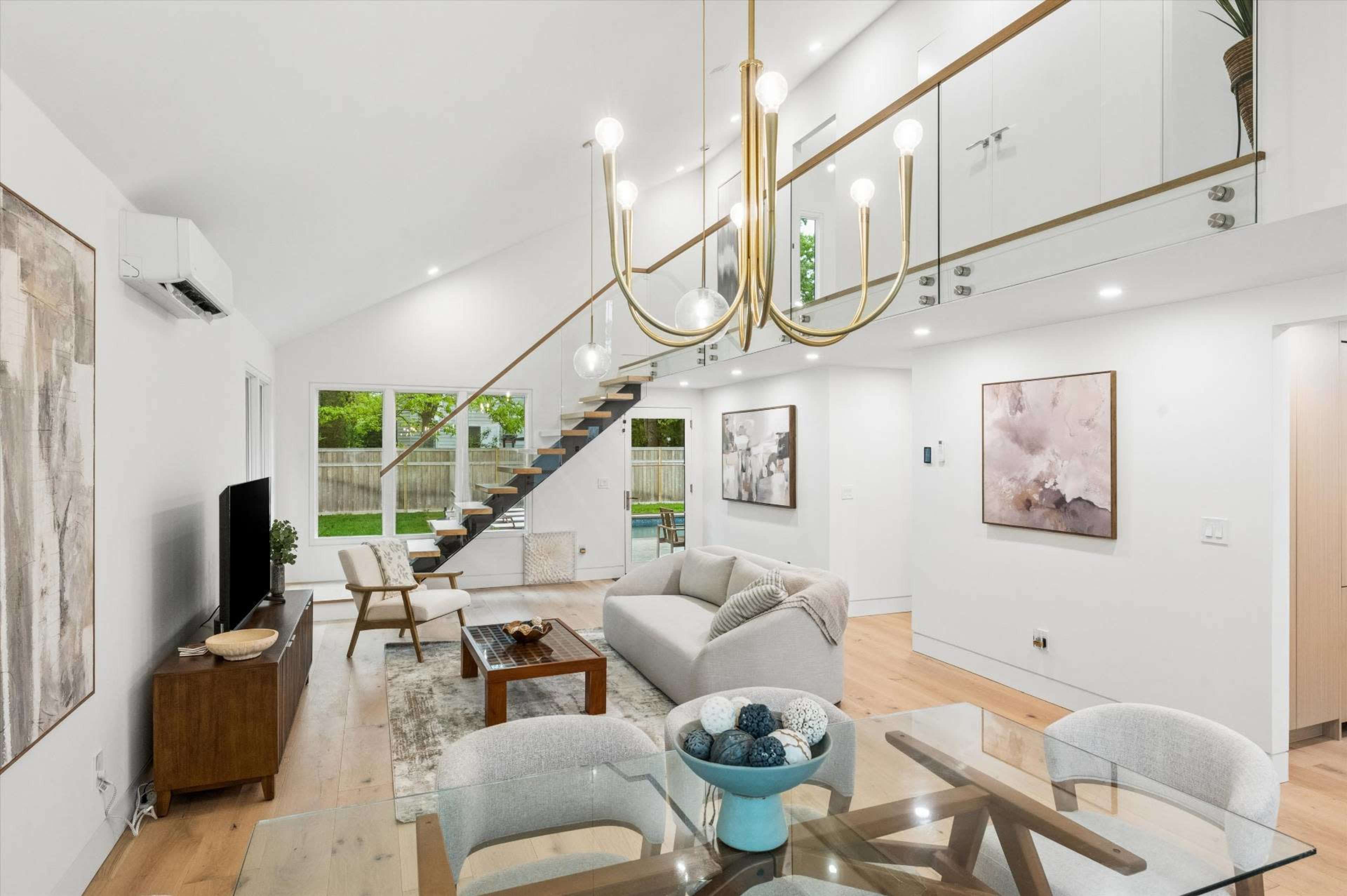 The image shows a modern living room with a staircase, a large window overlooking a yard, a TV stand, and minimalistic furniture.