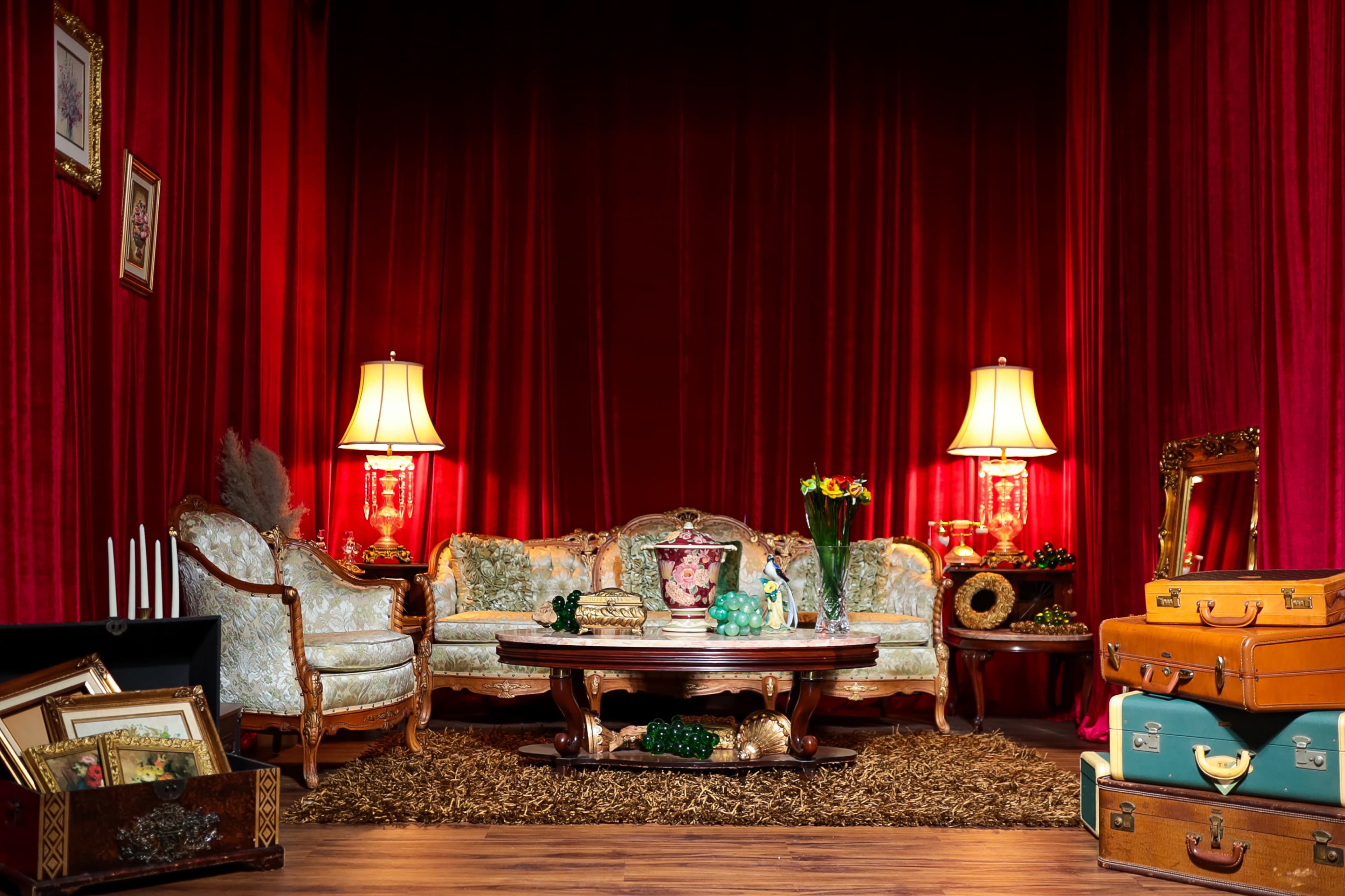 A vintage living room setting features a floral-patterned couch and armchairs, a wooden coffee table, two lamps, and various decorative objects, all set against a backdrop of red curtains.