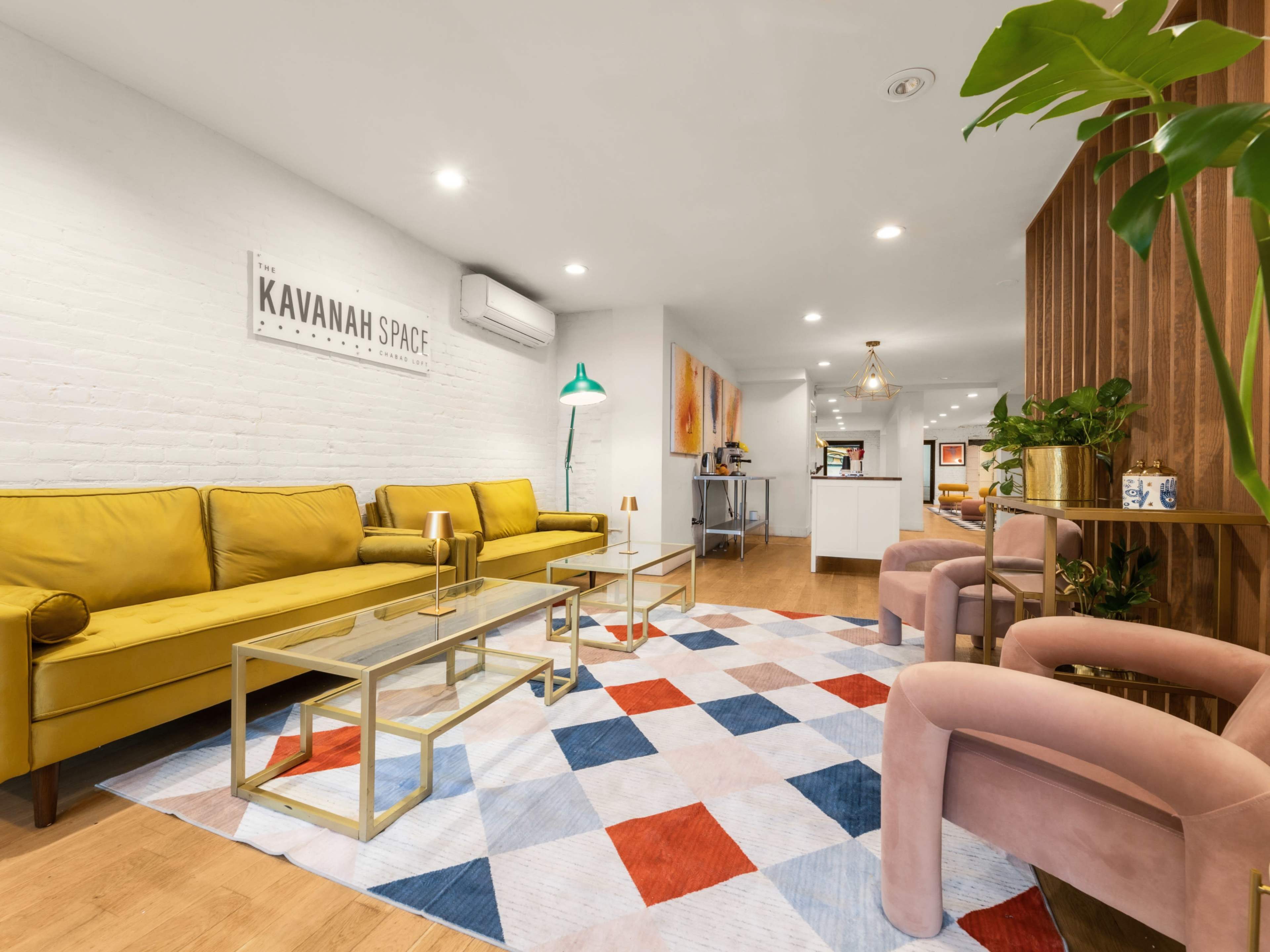 The image depicts a modern, inviting lounge area with yellow sofas, a colorful geometric rug, and decorative plants.