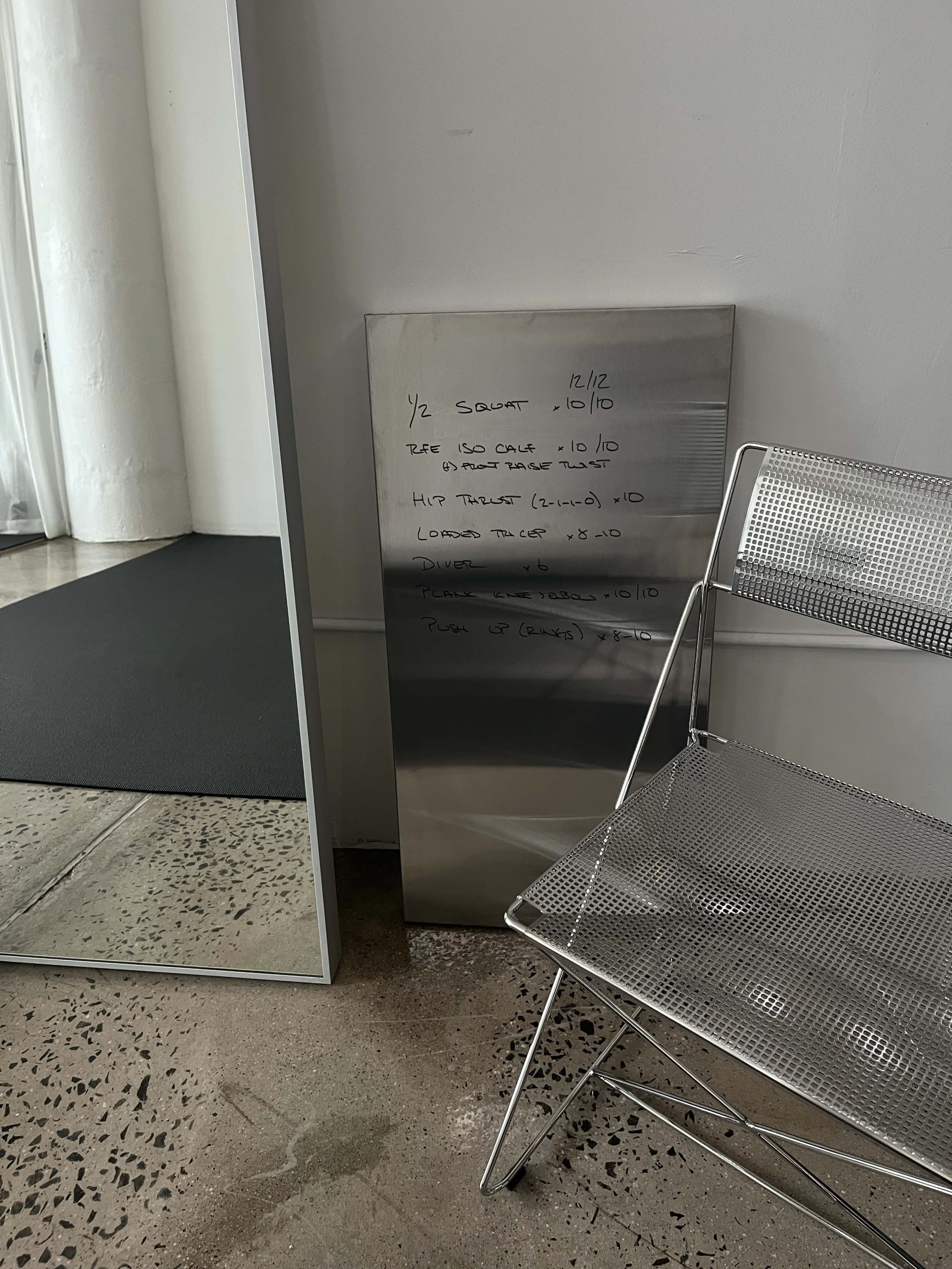 A stainless steel board with handwritten workout notes leans against a mirror beside a metal chair on a concrete floor.
