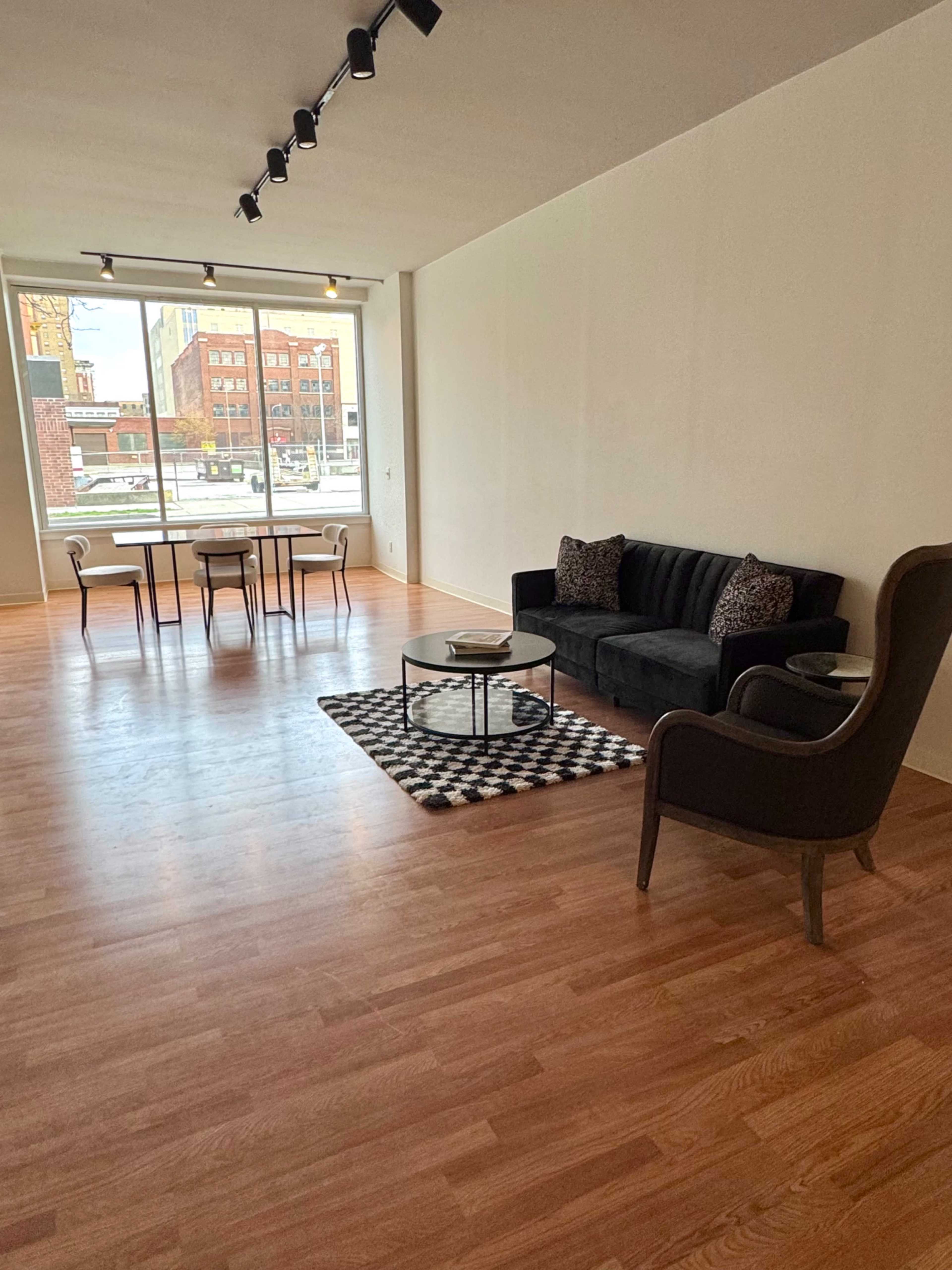 The image shows a minimalistic living space featuring a black couch, a round glass coffee table, a patterned area rug, a dining table with chairs, and large windows that let in natural light.