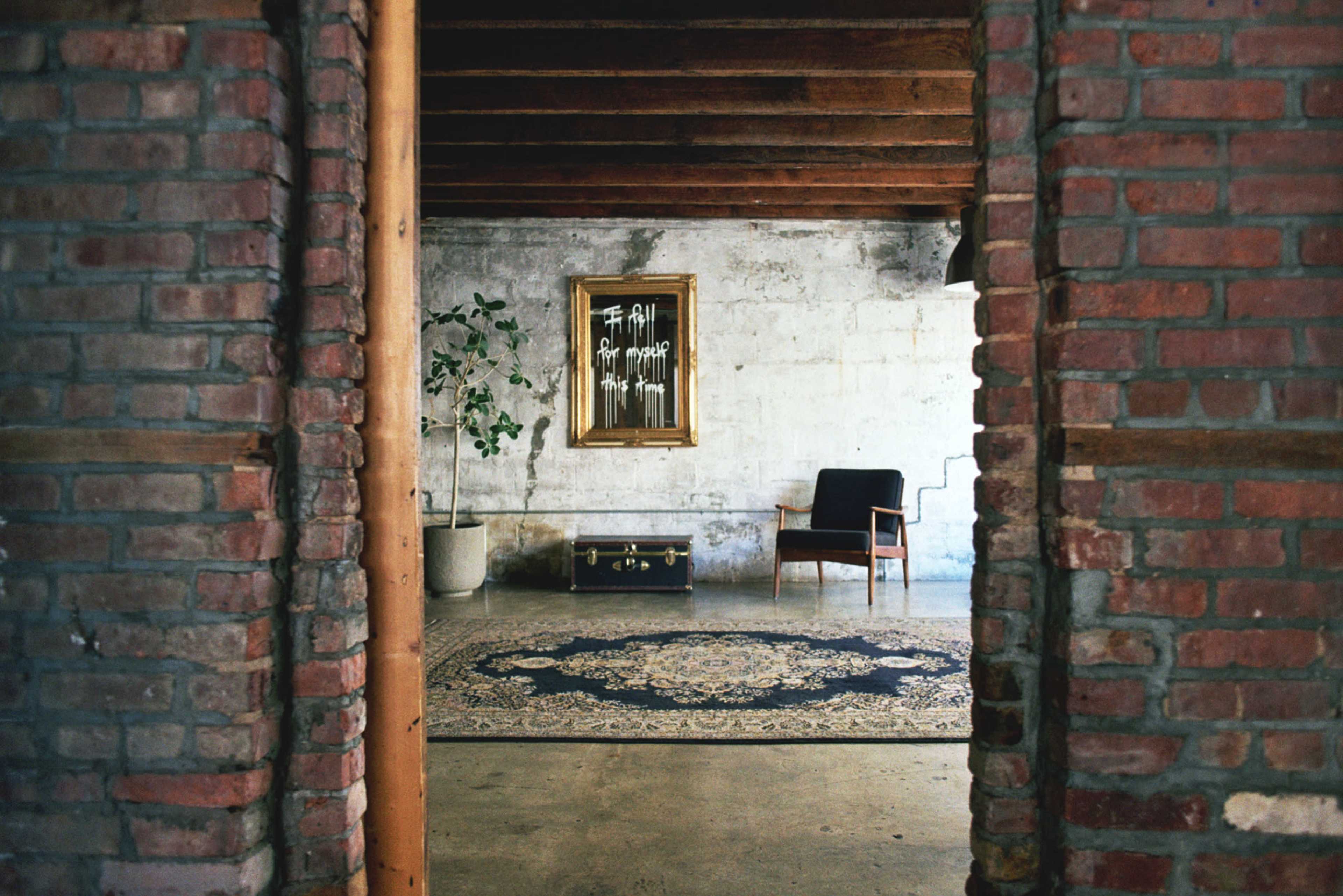 The image shows a brick archway framing a minimalist interior with a patterned rug, a potted plant, a vintage chair, and a mirror on the wall.