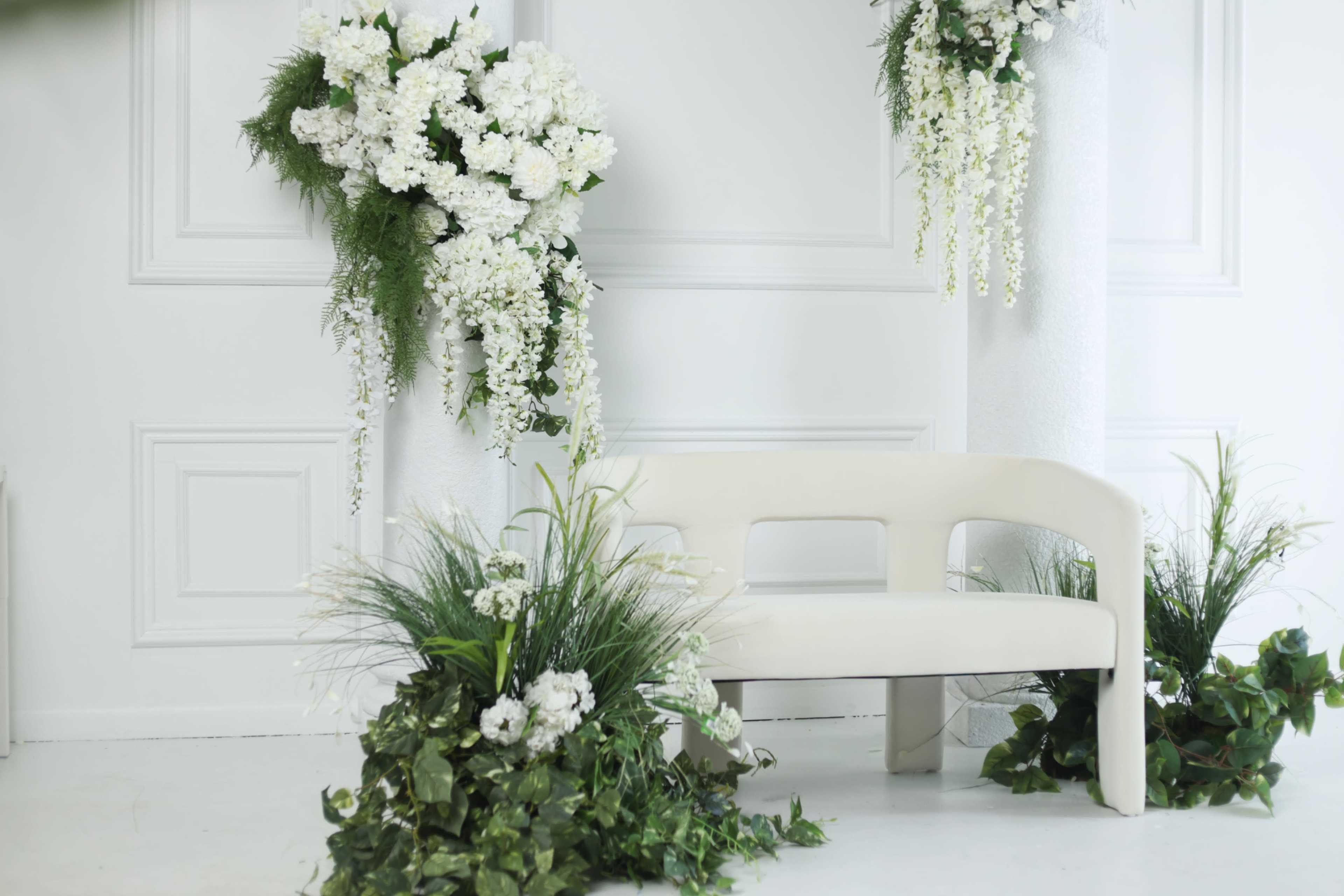 The image features a simple white bench surrounded by arrangements of white flowers and greenery against a plain backdrop.