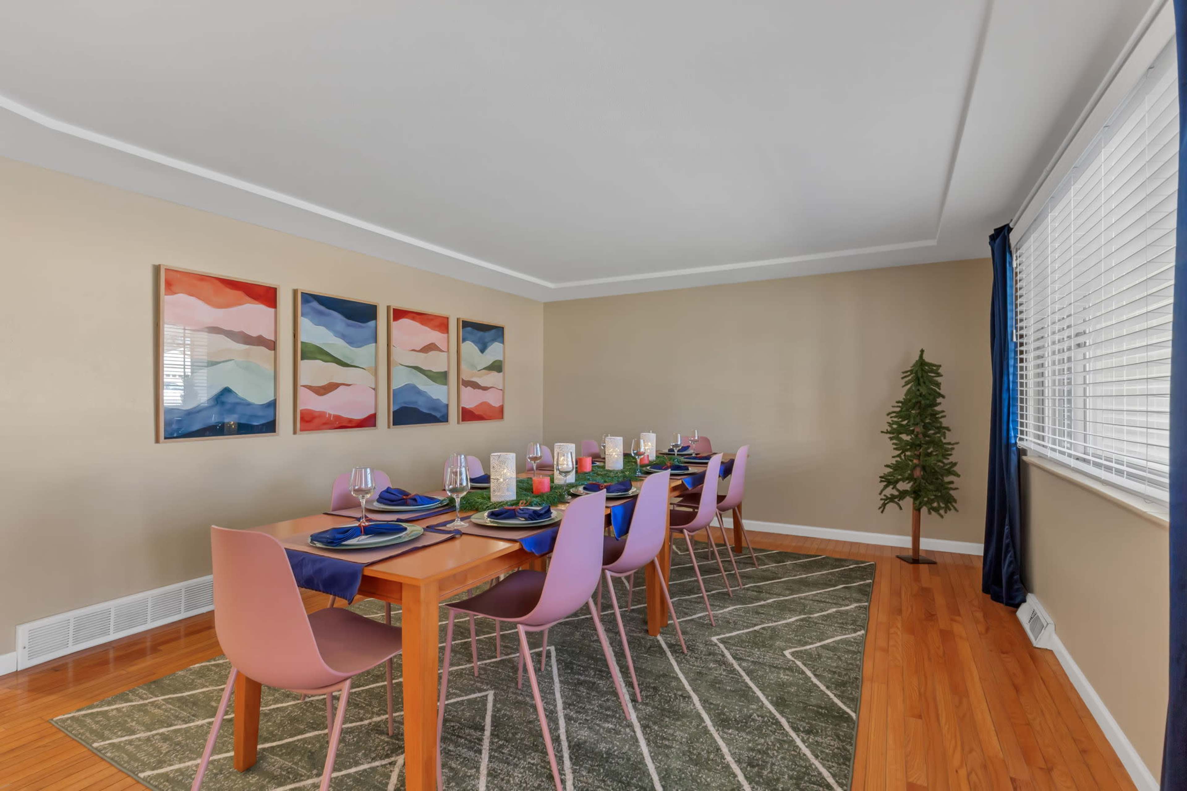 A dining room features a long wooden table set with plates and glasses, surrounded by pink chairs, and adorned with colorful wall art and a small tree in the corner.