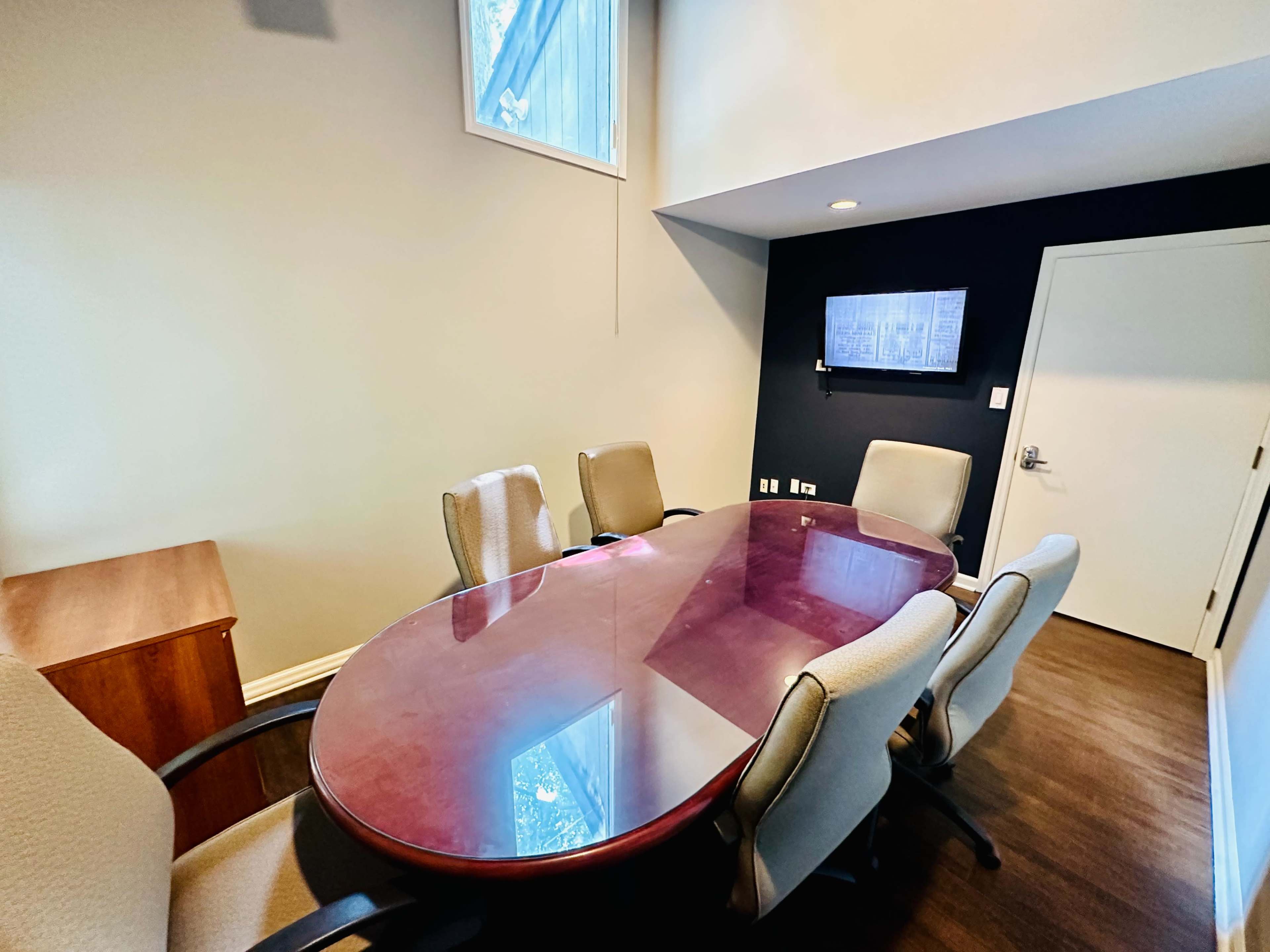 A conference room features an oval table surrounded by six chairs, with a wall-mounted screen and a window allowing natural light.